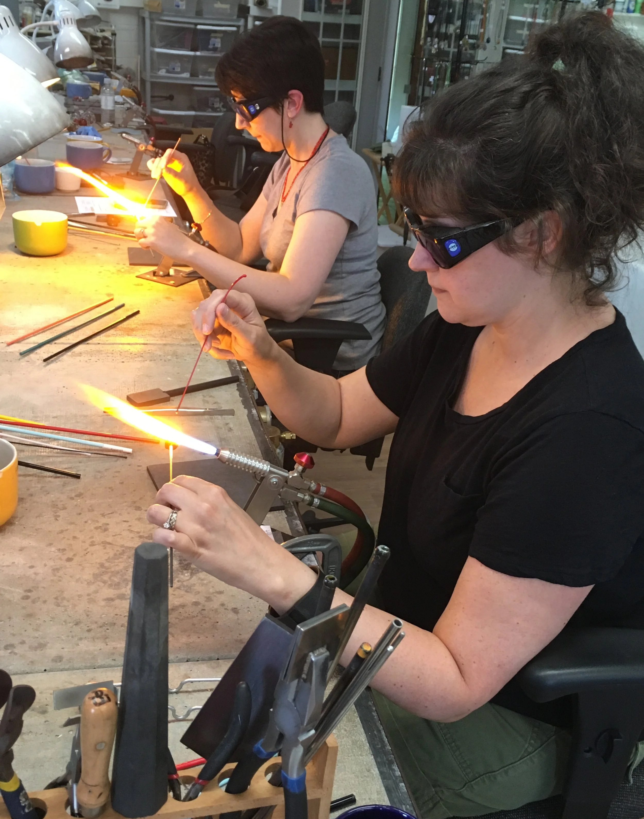 Two women working on glassblowing at a workbench in a workshop. They wear safety glasses and are using tools to shape molten glass with a torch. Various glassworking tools and supplies are on the table.