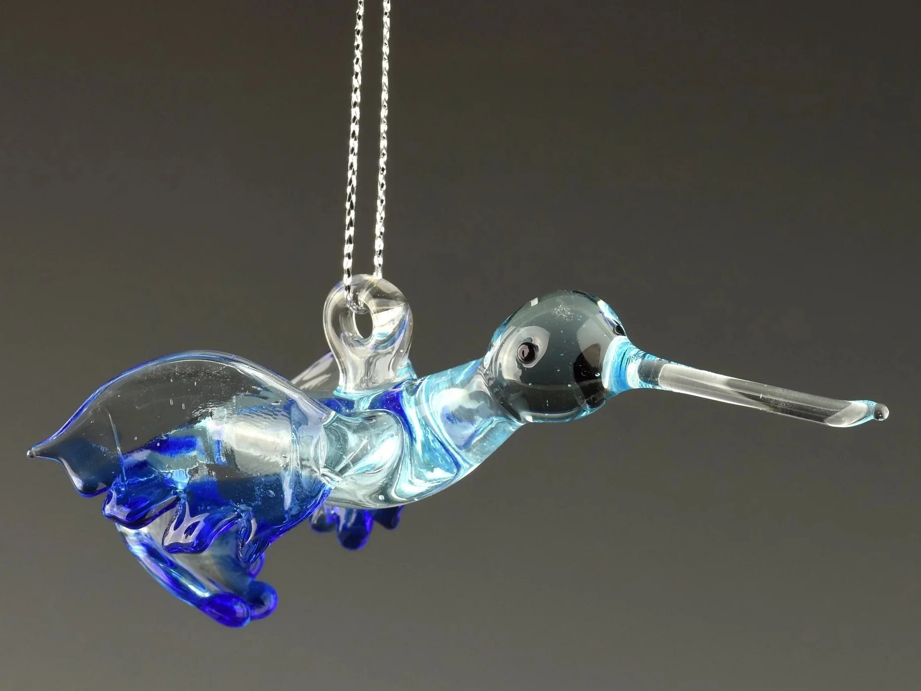 Close-up of a glass art piece, depicting a stylized bird with a long beak, made of clear and blue glass, hanging by a chain against a dark background.