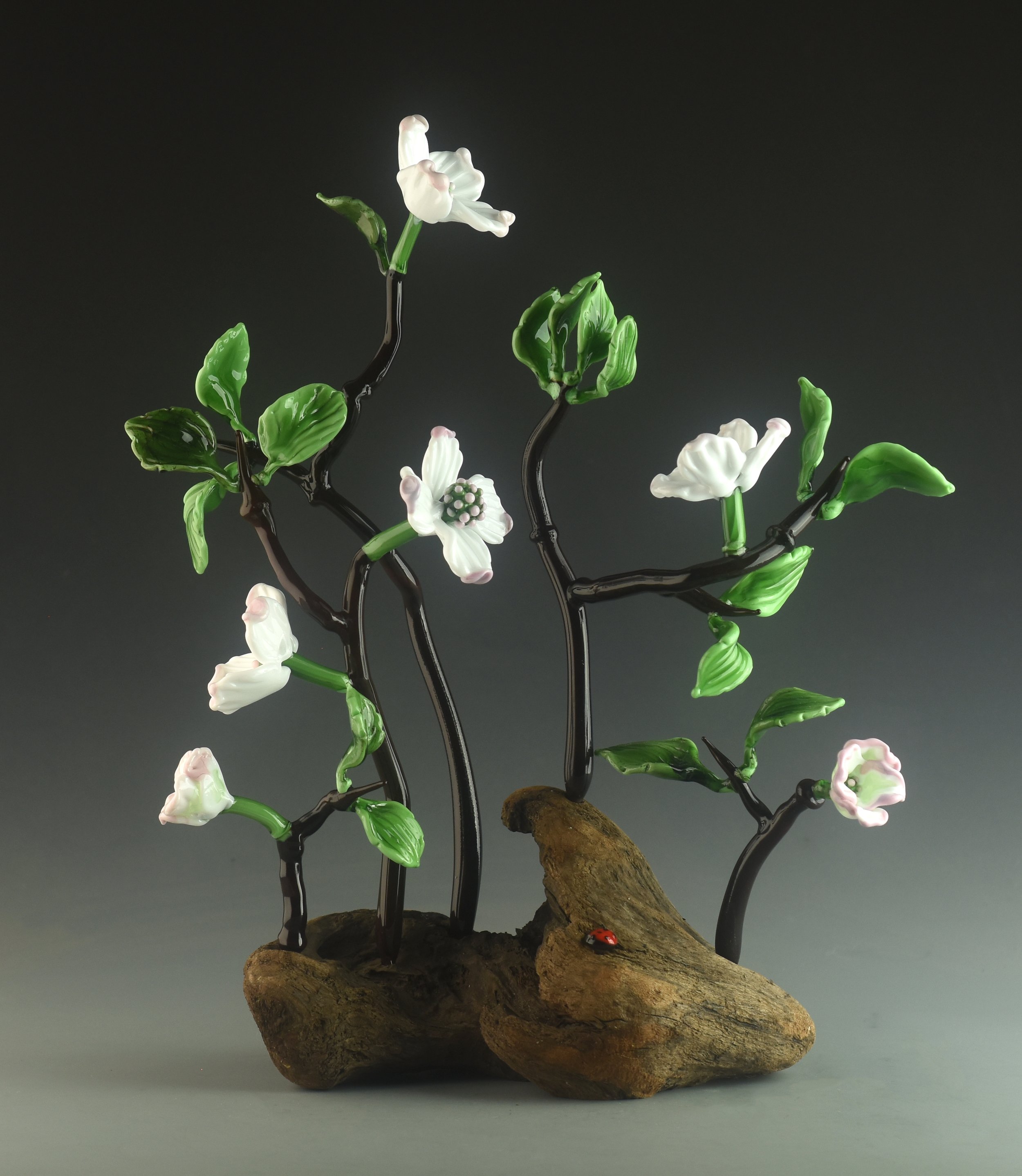 A decorative sculpture of a flowering plant made of glass, with white blossoms, green leaves, and dark, twisting branches on a piece of wood with a tic-tac-toe ladybug at the base, set against a dark background.
