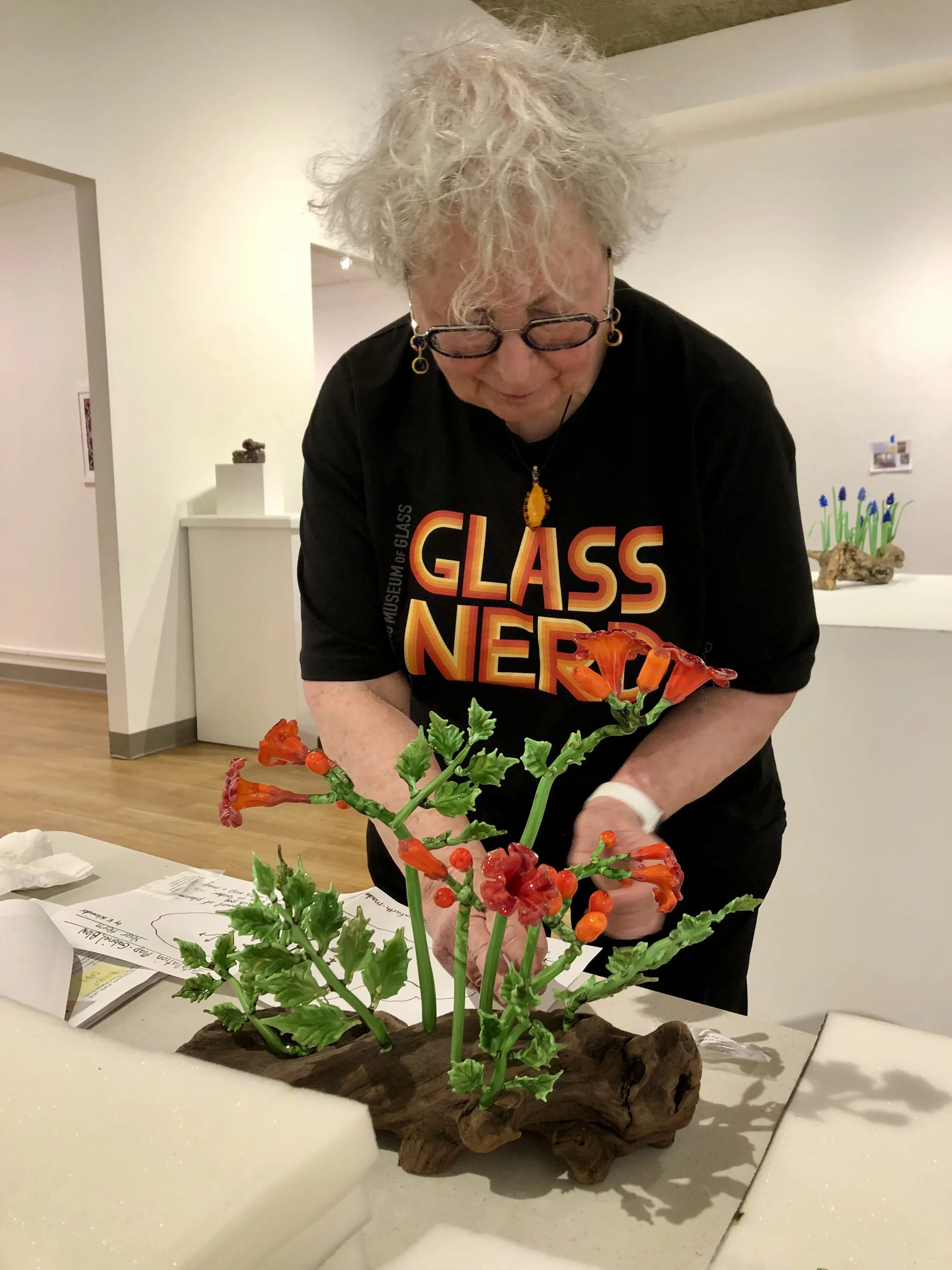 An older woman with gray, curly hair and glasses, wearing a black shirt with colorful text, is working on a glass art project featuring orange and red flowers with green leaves, placed on a piece of driftwood. She is indoors with white walls and some framed pictures in the background.