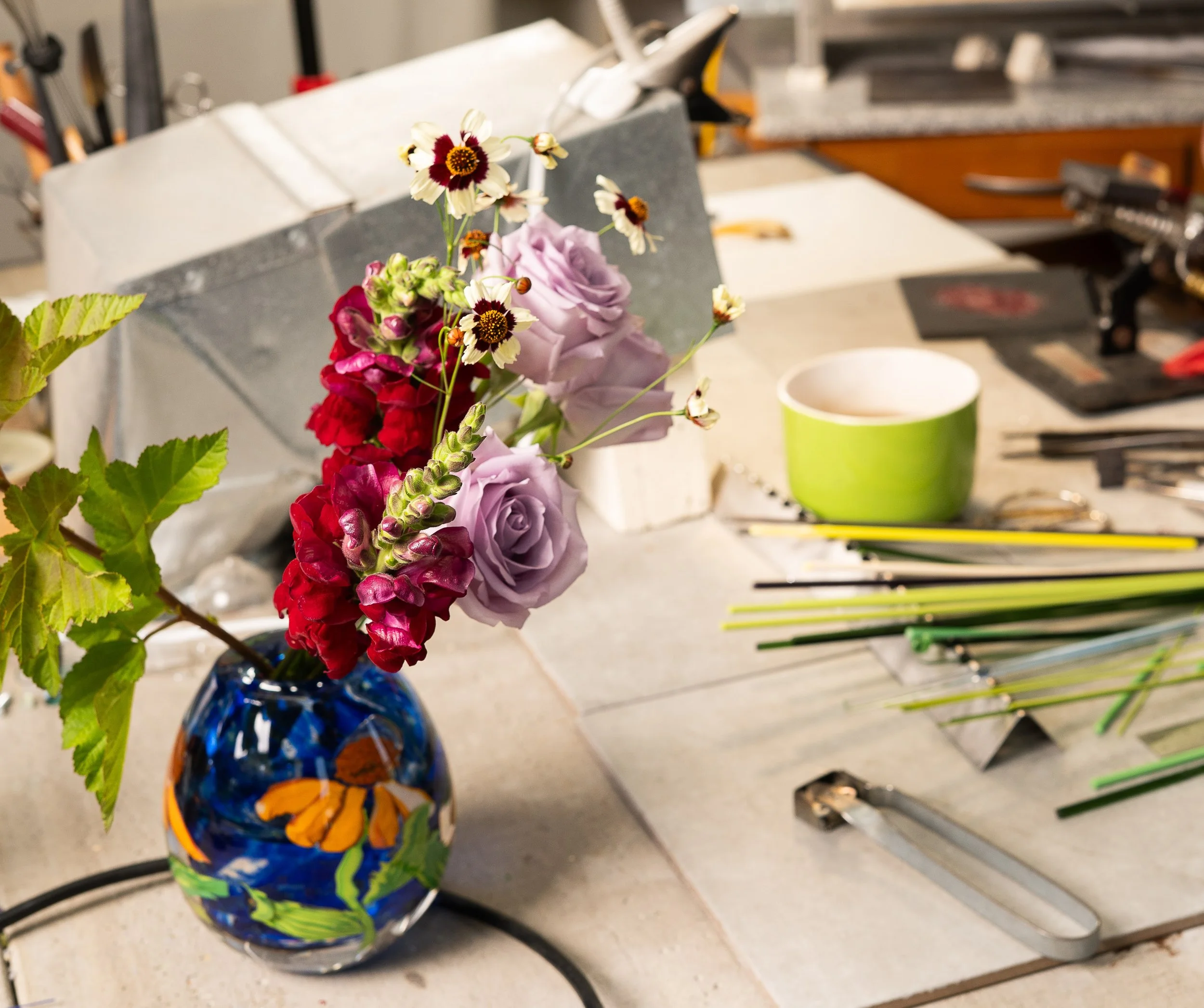 A colorful floral arrangement in a blue, orange, and yellow painted vase on a kitchen countertop. The bouquet includes purple roses, red flowers, and white flowers with dark centers, with some green foliage visible.