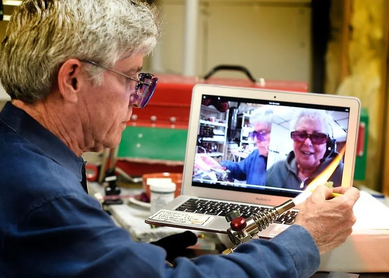 An elderly man with gray hair and glasses working in a workshop, video calling on a laptop with two women on the screen, one of whom is smiling and wearing sunglasses.