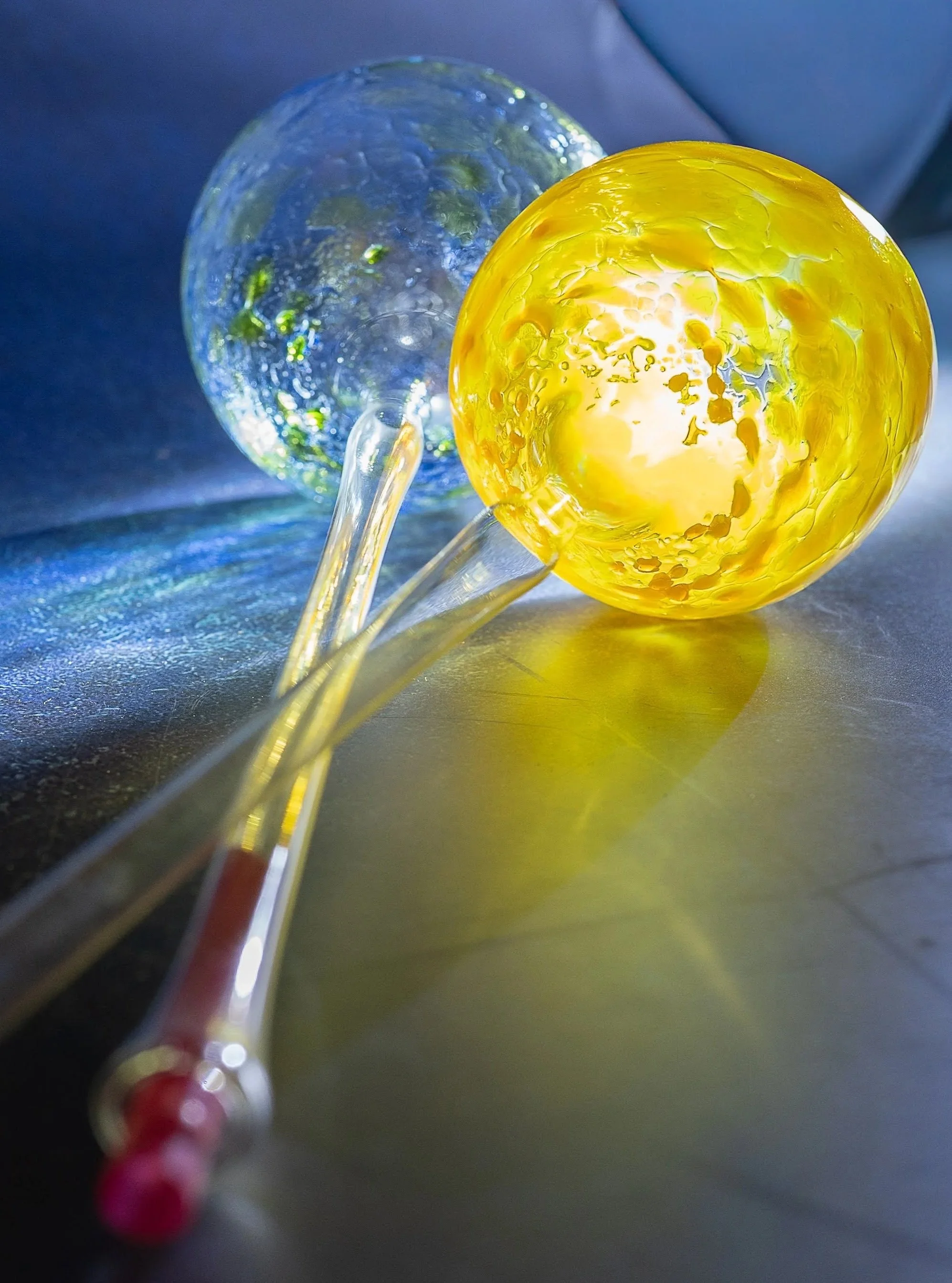 Close-up of two glass water globes, one yellow and one clear with green and blue swirls, with a reflective surface underneath.