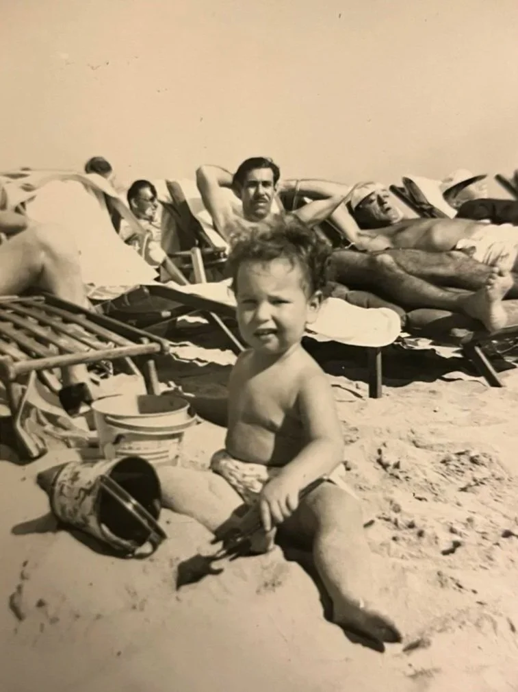 A young boy at the beach sitting on the sand, holding a small shovel and toy, with beach chairs and people relaxing in the background.