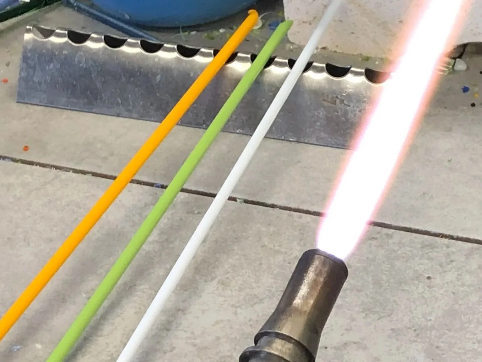 A welding torch emitting a bright light aimed at four colored pipes (yellow, green, white, and a third color not visible) on a concrete surface, with a metal bracket or support nearby.