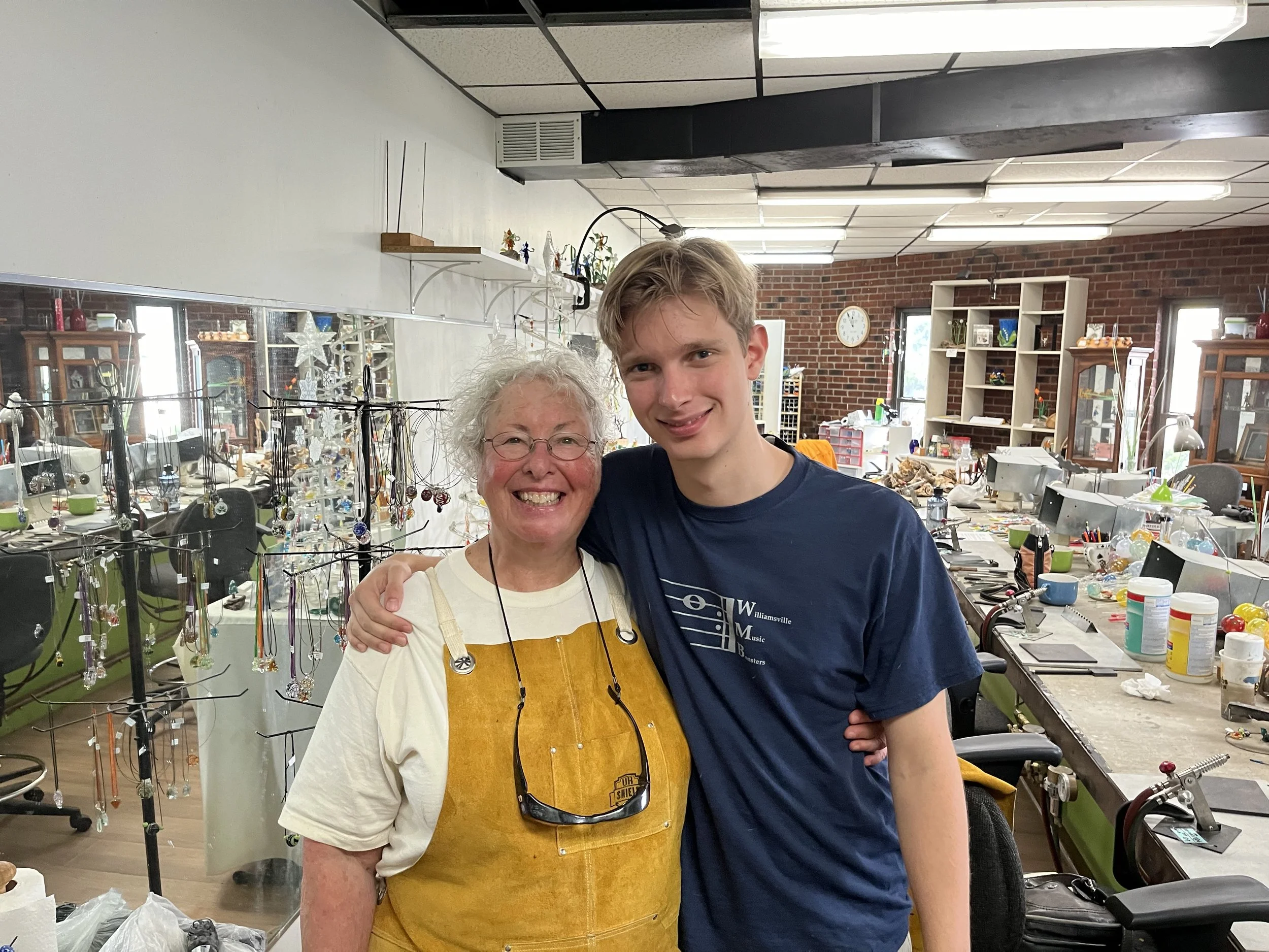 Two people smiling and hugging in a jewelry workshop. The woman on the left has curly white hair, glasses, and is wearing a white shirt. The young man on the right has short blonde hair, is wearing a navy blue t-shirt, and has braces.