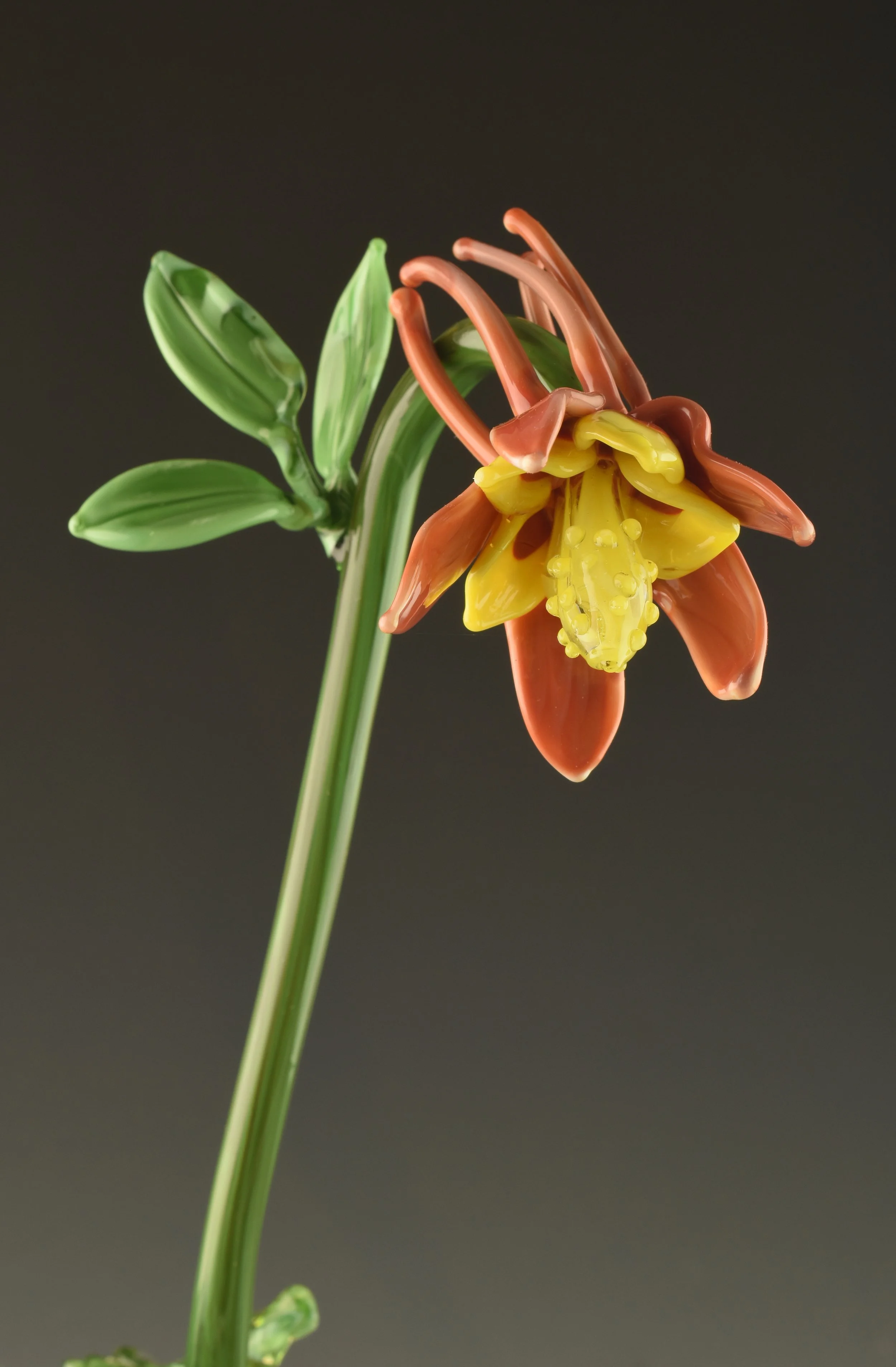 A close-up of a glass flower sculpture resembling a lily with green leaves, orange petals, and a yellow center against a dark background.