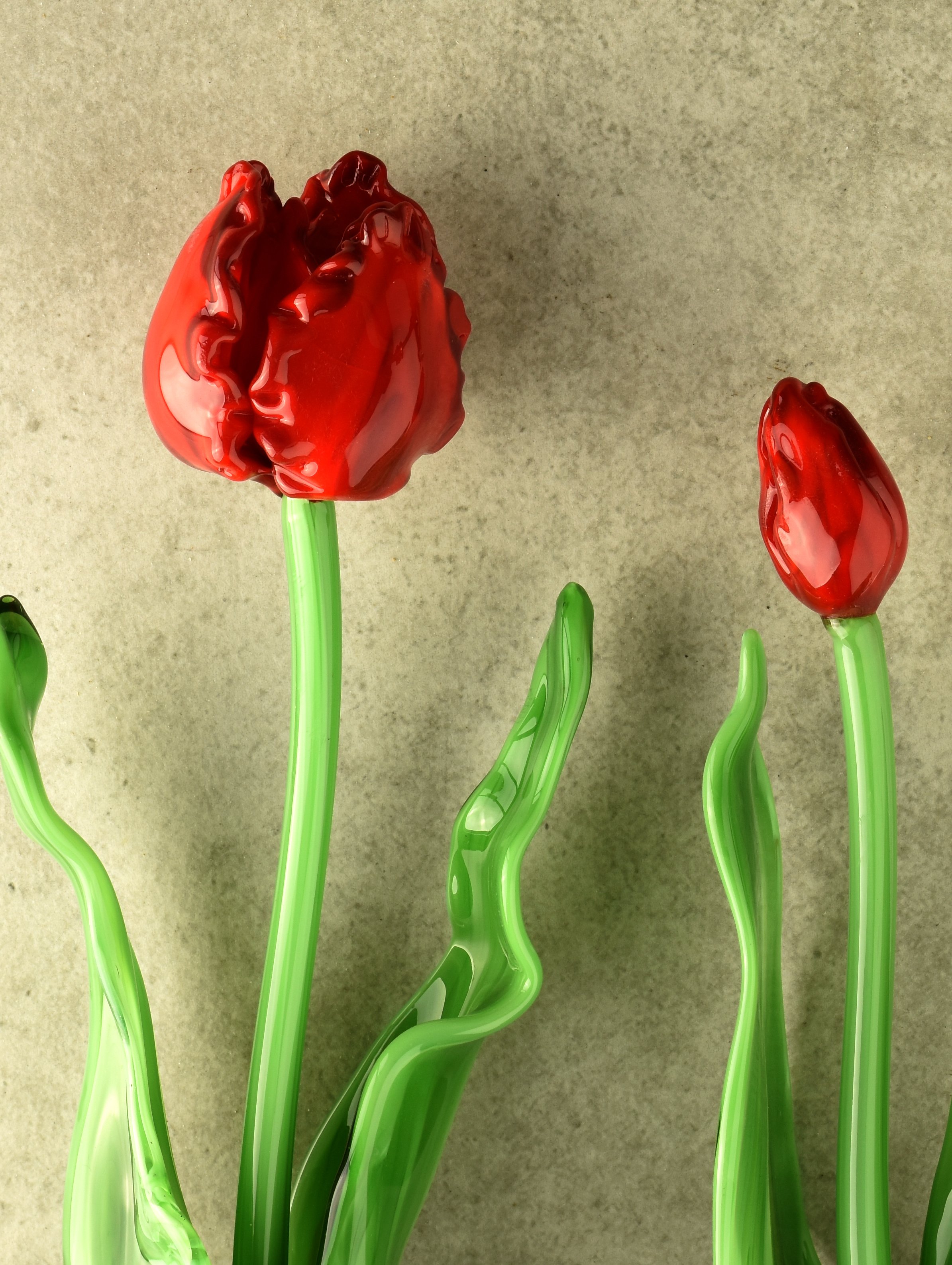 Glass art flowers with red blooms and green stems and leaves, set against a plain background.