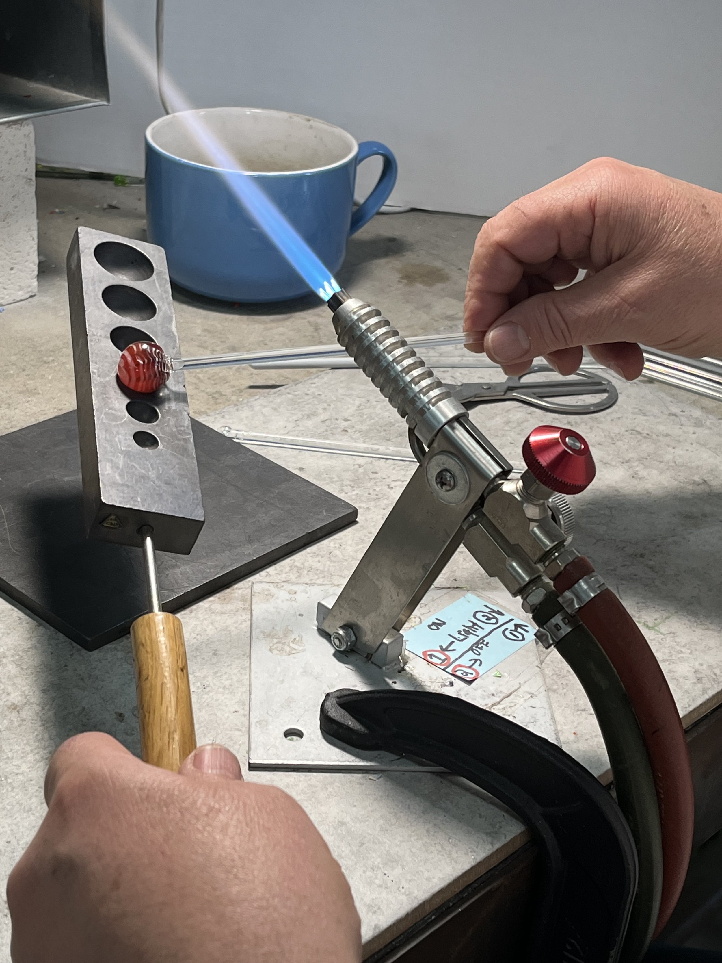 A person holding a glass stick near a metal stand with a red and white object, a blue mug in the background, and a blue flame from a torch aimed at the object.
