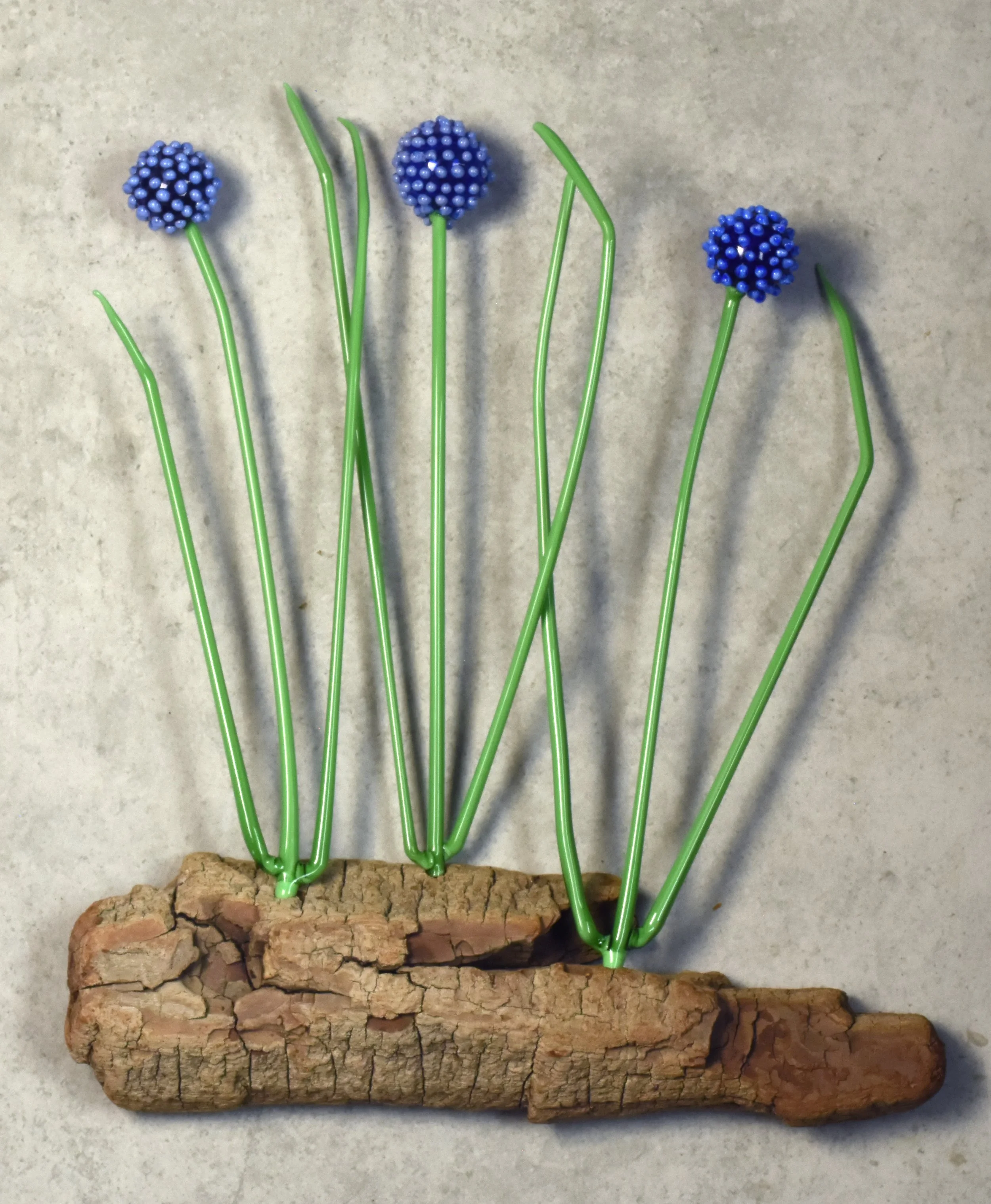 Artificial blue flowers with green stems stuck into a piece of cork on a gray surface.