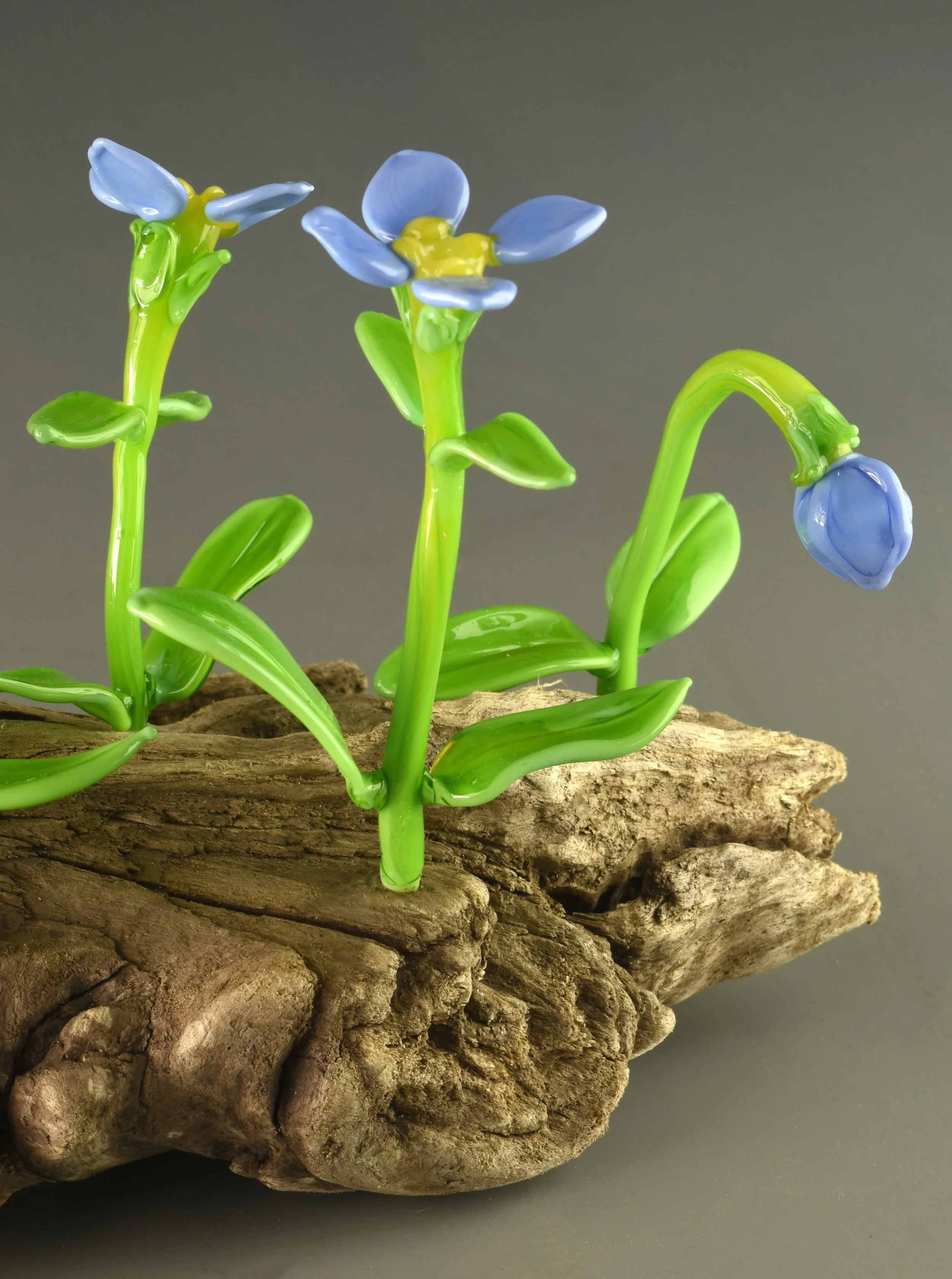 A glass art flower arrangement with three blue flowers and green leaves on a piece of wood