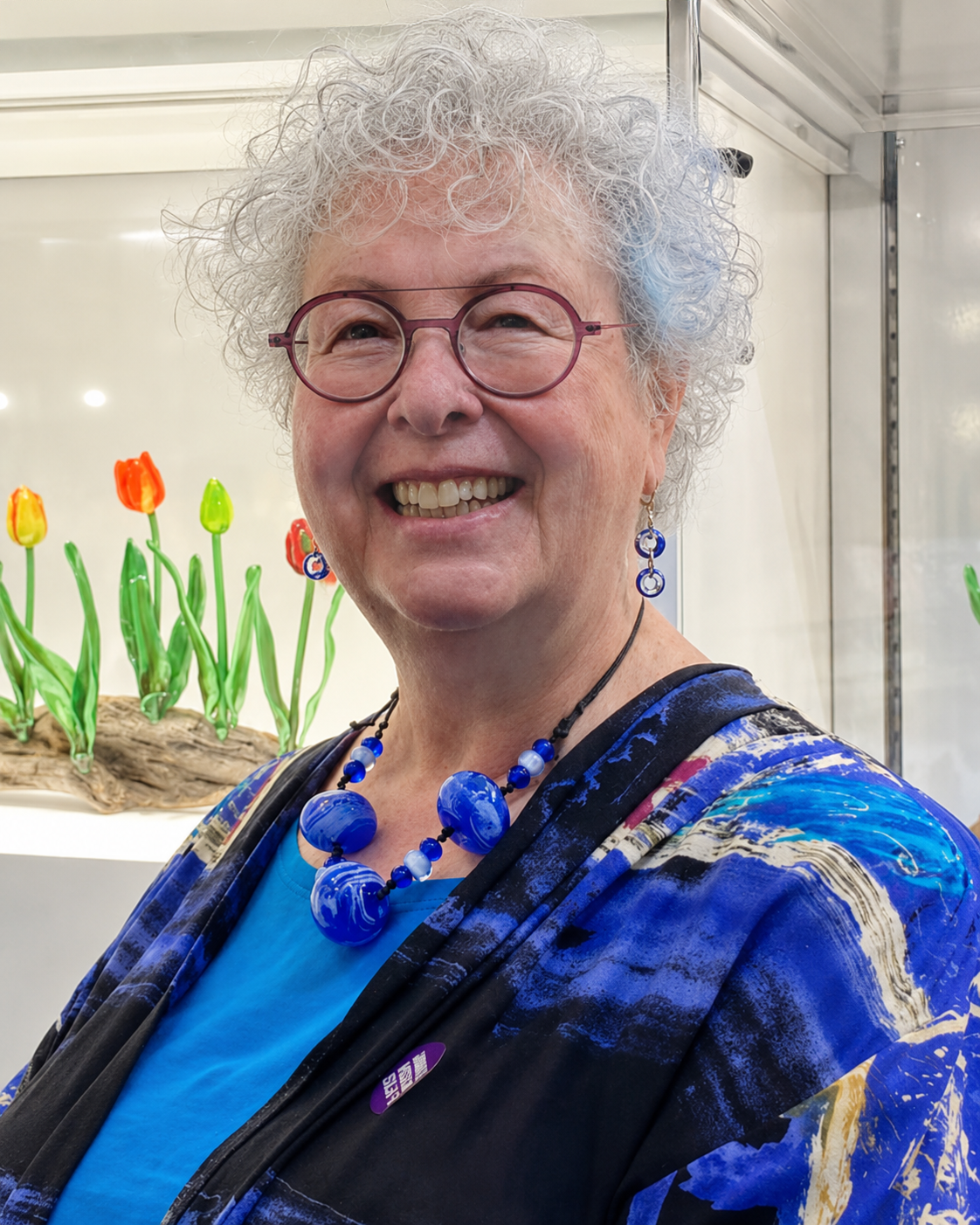 A smiling glass artist with blue jewelry, and a colorful patterned jacket, standing in front of glass art of tulips.