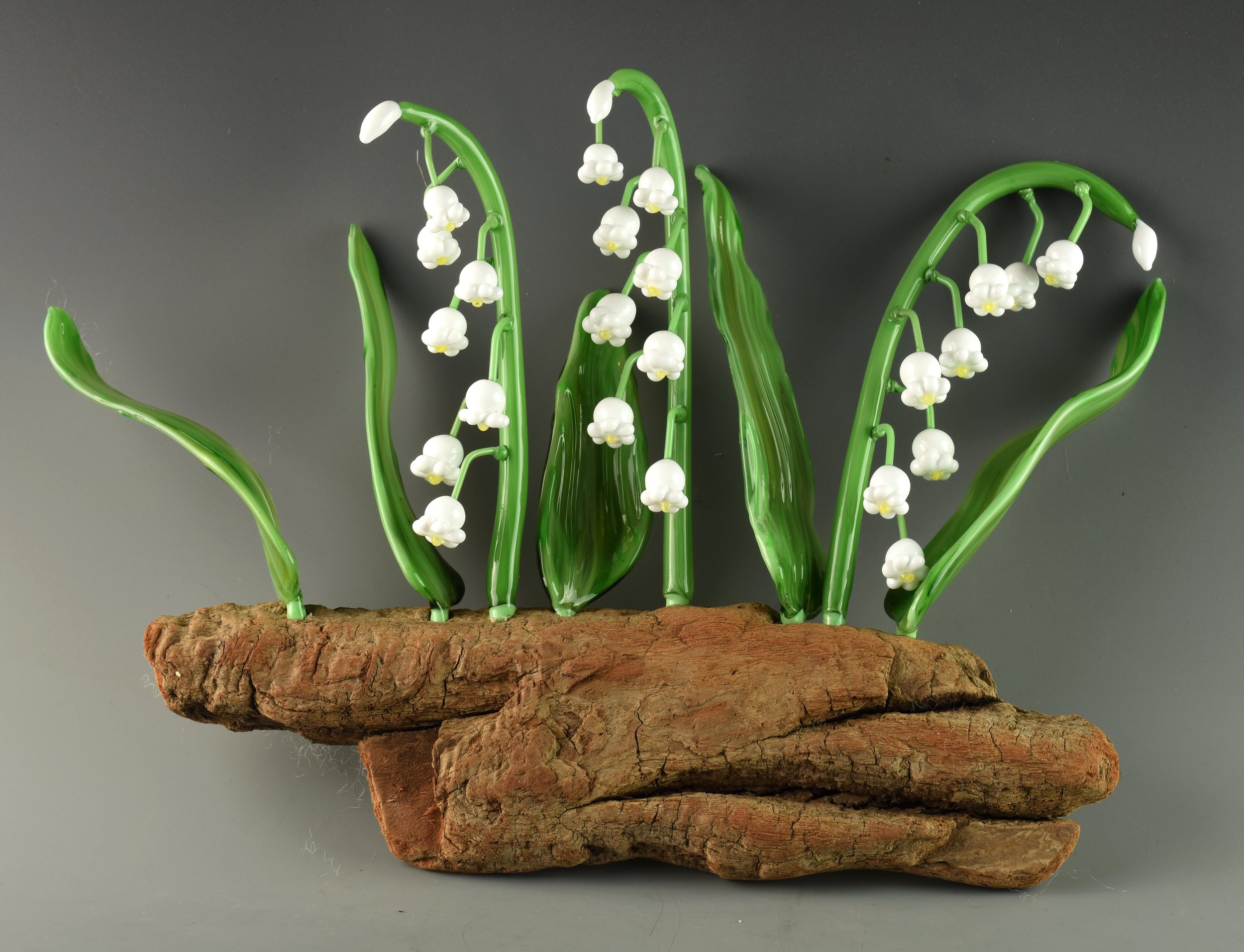 Glass sculpture resembling lily of the valley flowers with green leaves on a piece of wood.