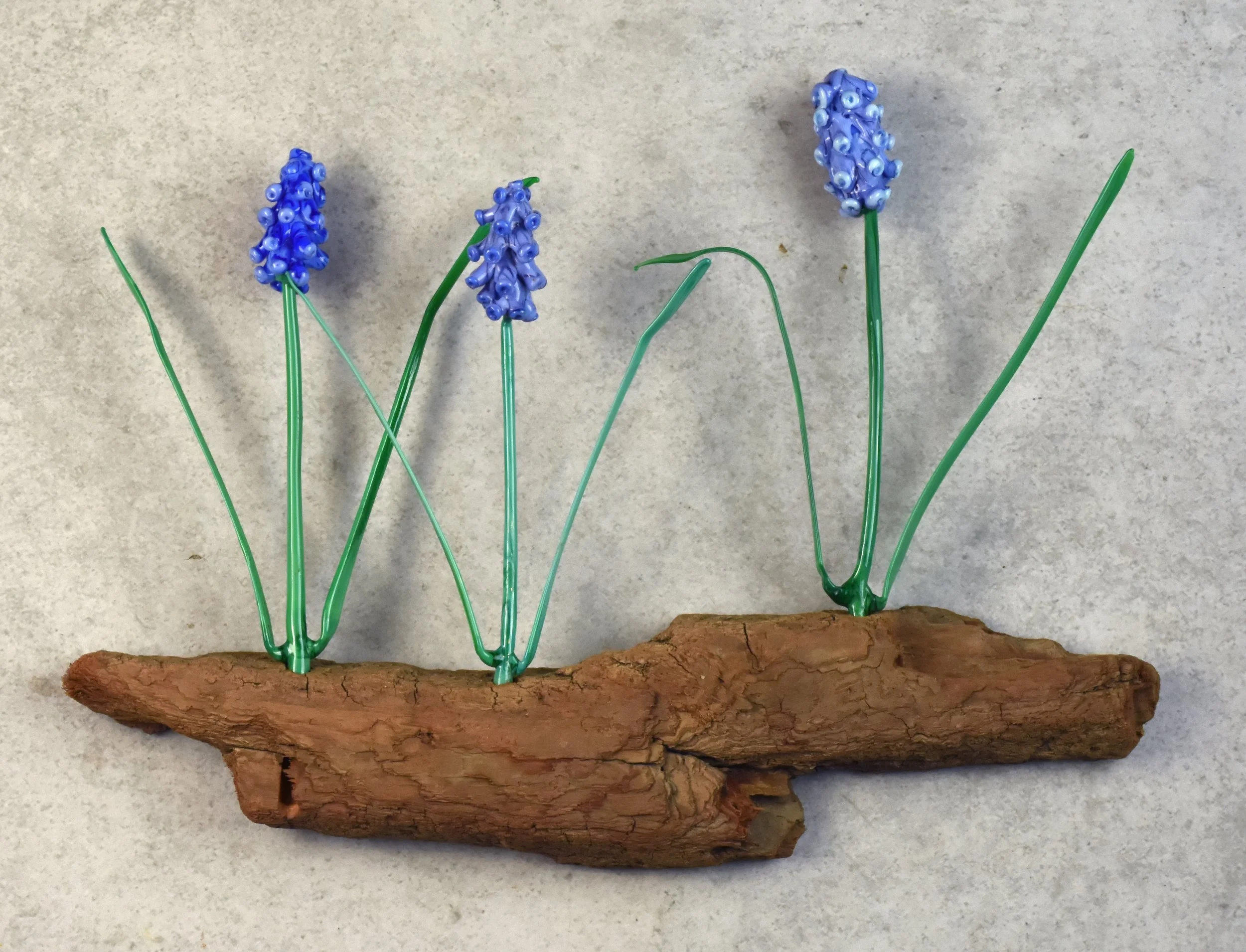 Artificial grape hyacinth flowers made of blue beads and green wire, attached to a piece of driftwood.