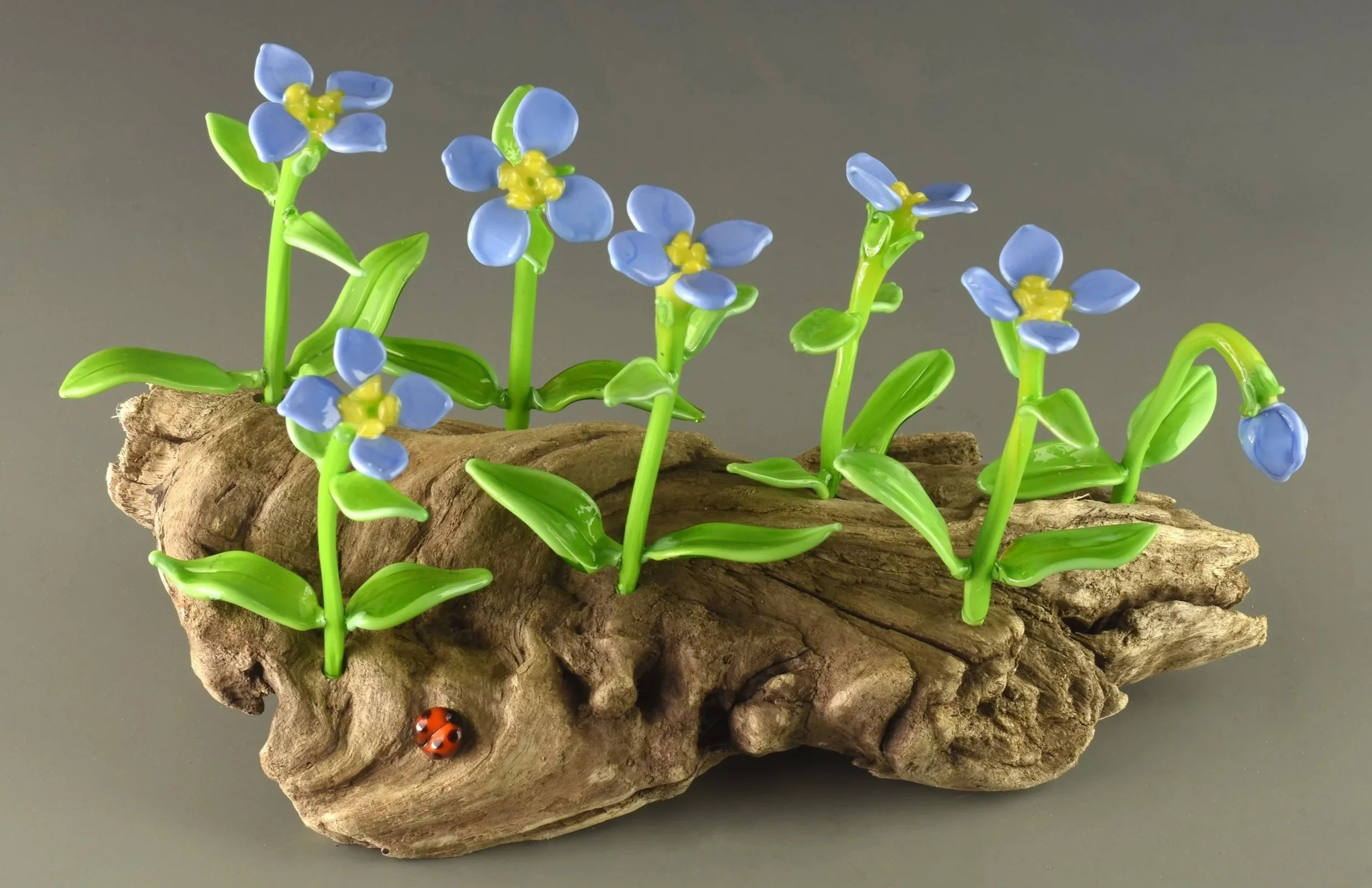 A decorative art piece resembling a small garden with blue and yellow glass flowers and green leaves, mounted on a textured brown wooden slab with a ladybug near the base.