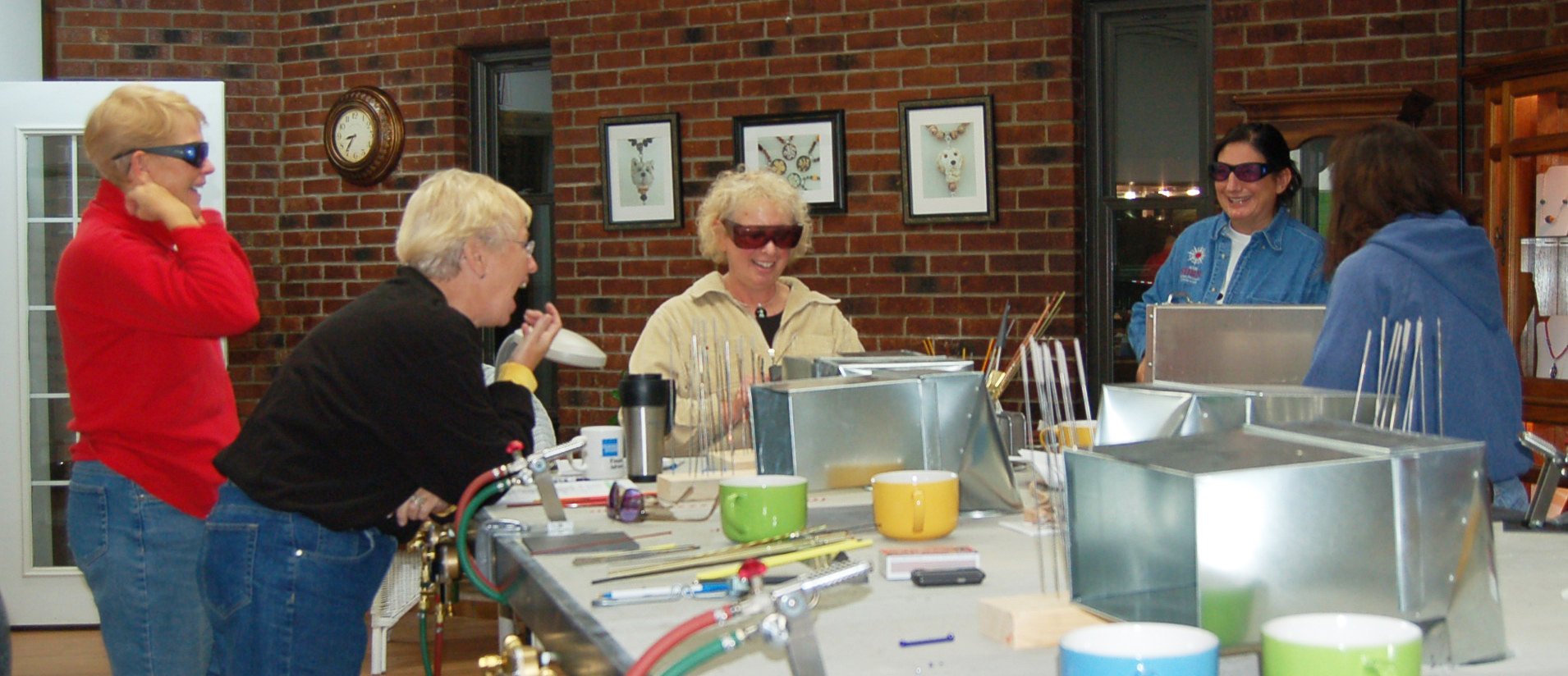 Five women working with glass in a workshop, some wearing protective glasses, surrounded by tools and colorful cups, with brick walls and framed artwork in the background.