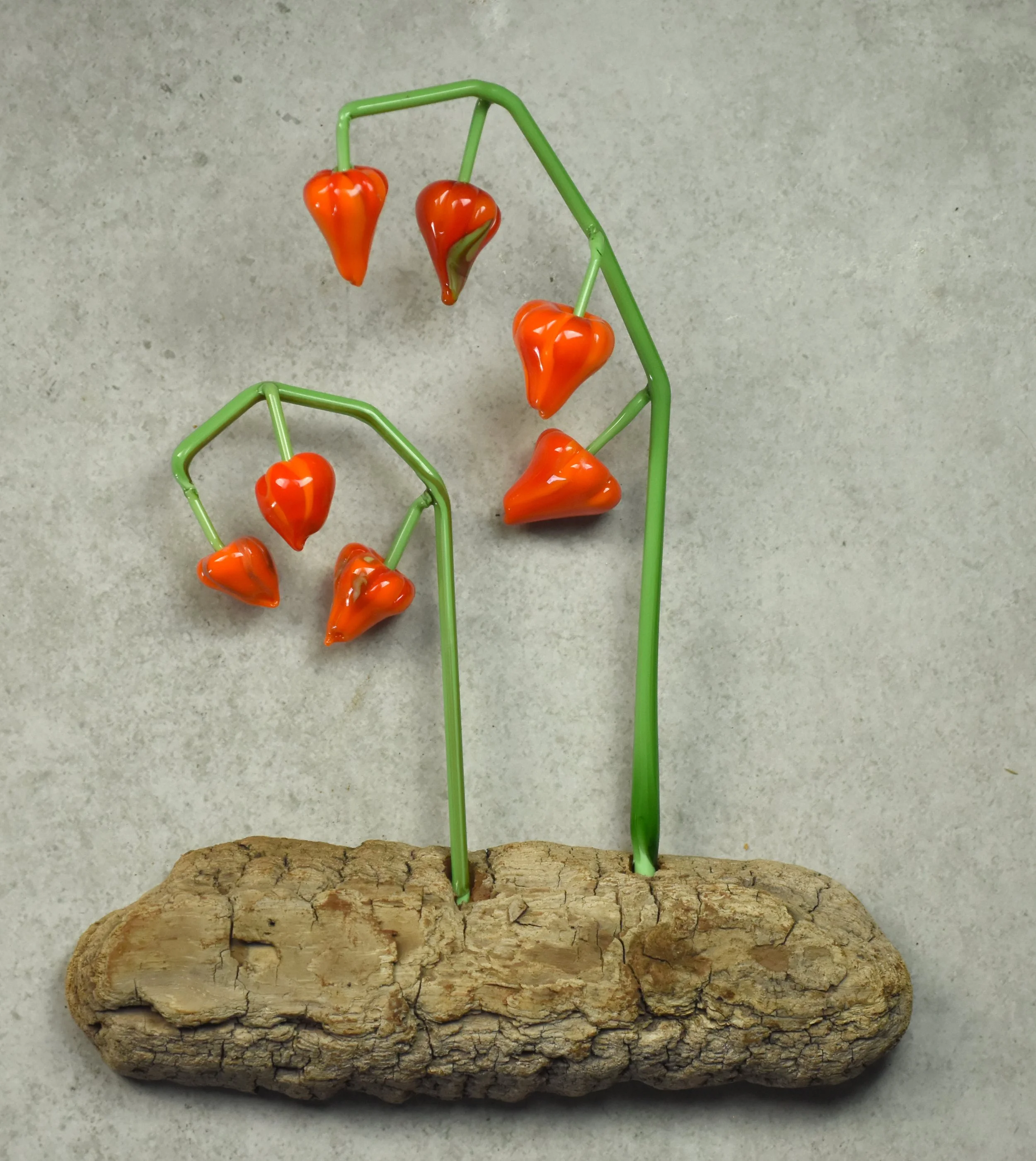 Colorful glass peppers attached to green wire stems, displayed on a piece of wood.