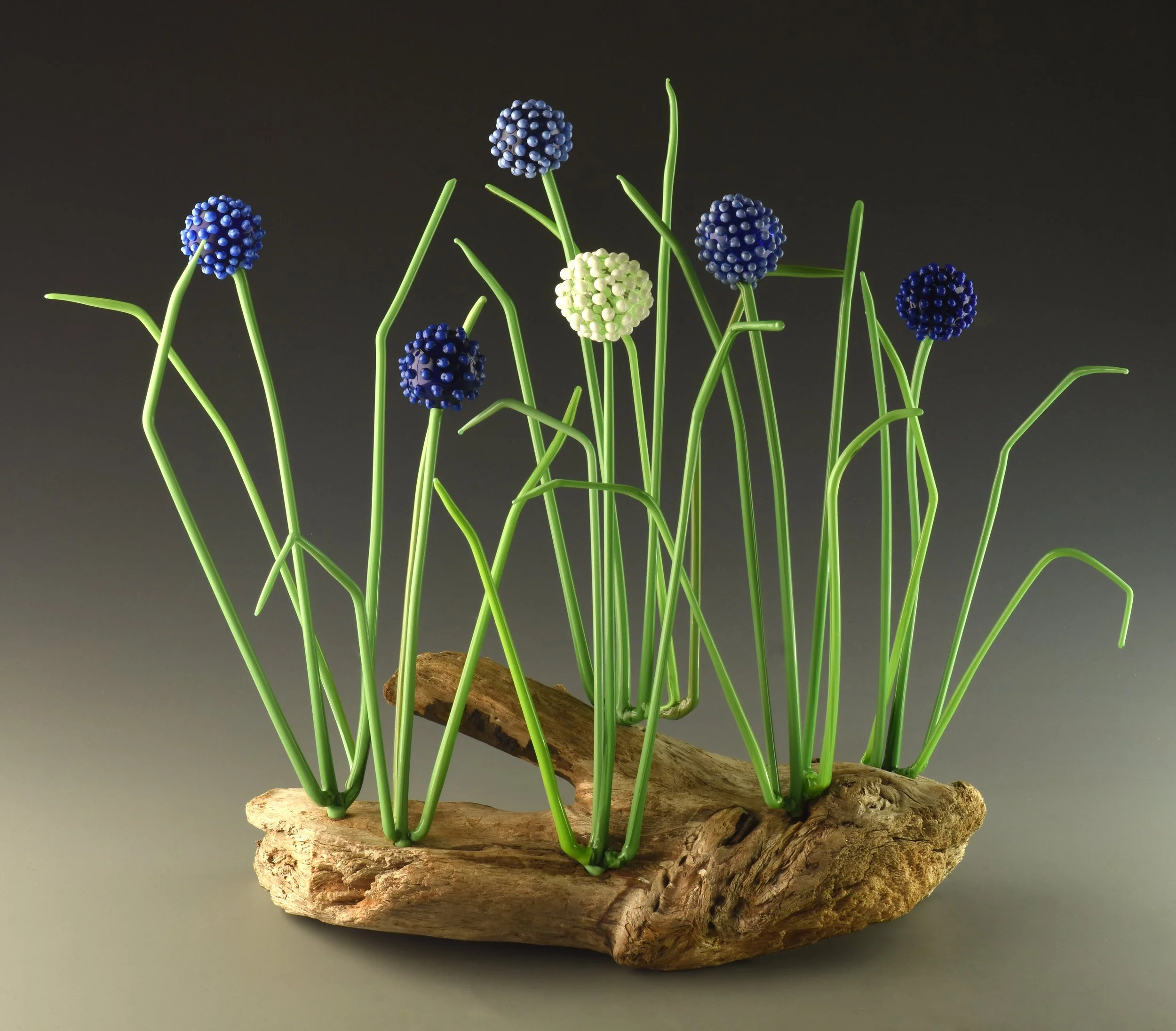 Artistic sculpture resembling a cluster of blue and white berries with green stems, mounted on a textured wooden base, set against a plain dark background.