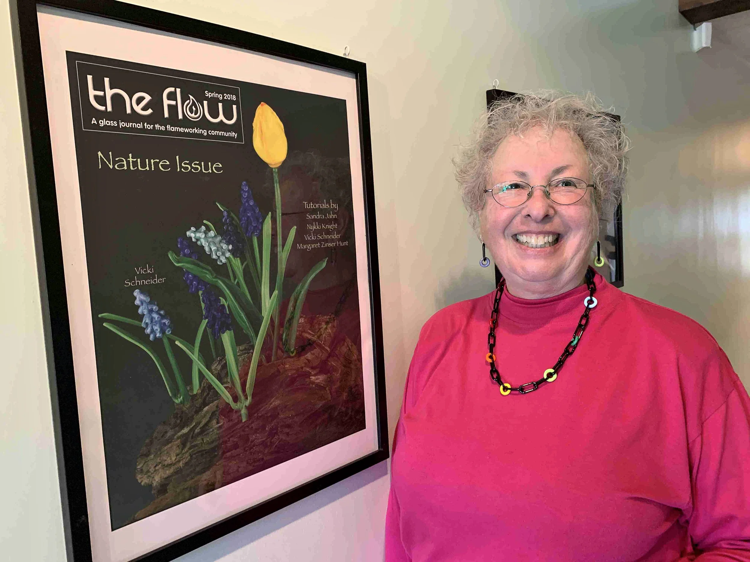 A woman with glasses and curly gray hair smiling while standing next to a framed magazine cover titled 'the flow' with a subtitle 'Spring 2018' and 'A glass journal for the flameworking community'. The cover features an illustration of a yellow flower with a green stem and purple blues, with a dark background.