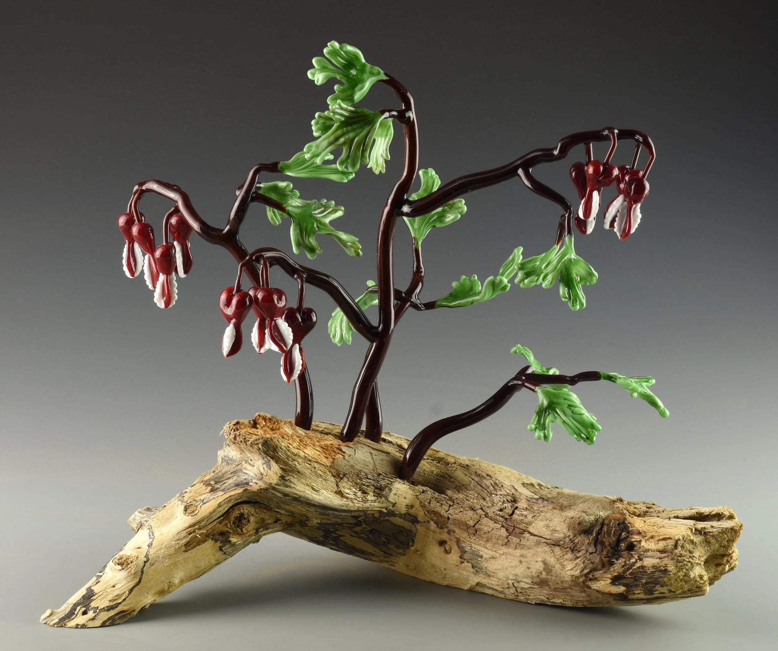 Glass sculpture of a tree with green leaves and red and white hanging flowers mounted on a piece of driftwood.