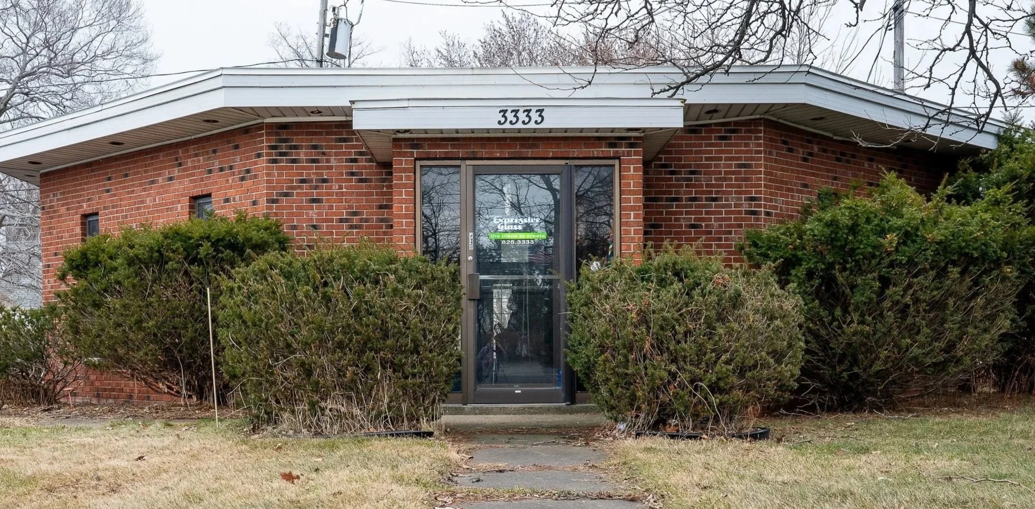 Red brick commercial building with a white overhang at 3333, surrounded by green bushes and a small front yard. The entrance has a glass door with a green sign, and the background shows a cloudy sky with leafless trees.