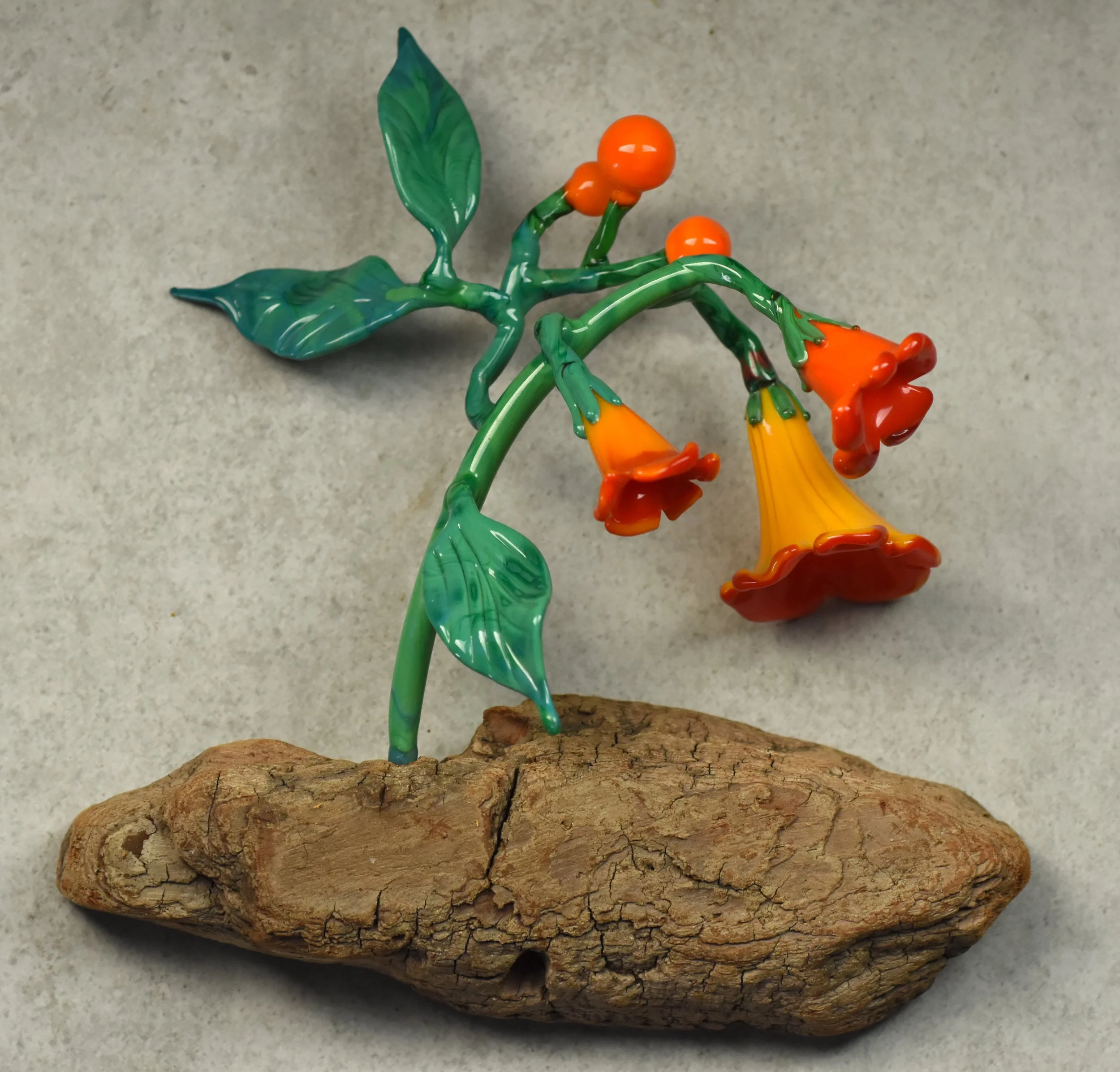 Colorful glass sculpture resembling a flower with green leaves, orange buds, and yellow petals, mounted on a piece of textured wood.