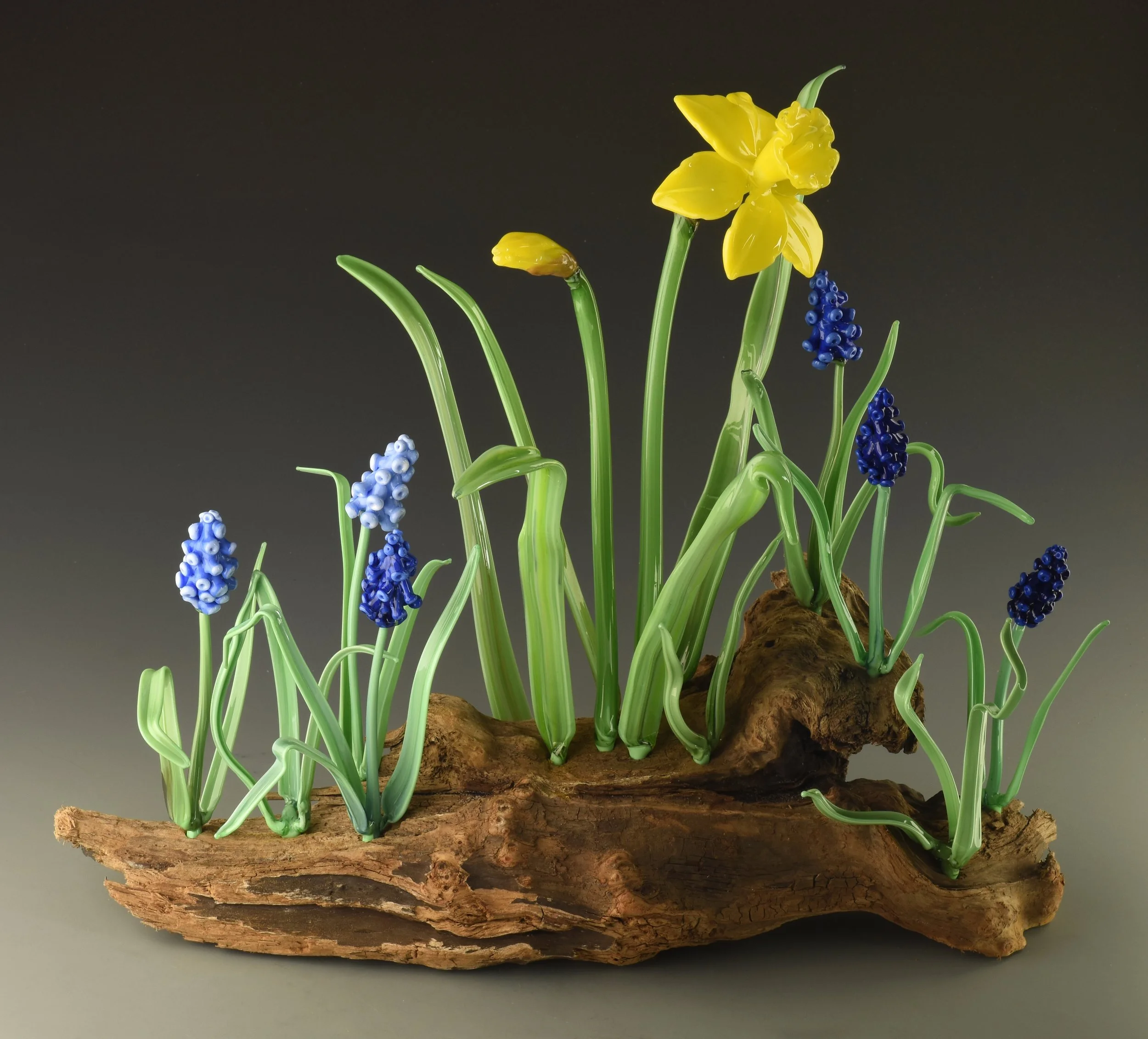 Glass sculpture of yellow daffodil flowers with green stems, blue grape hyacinths, and moss on a large piece of driftwood against a plain dark background.