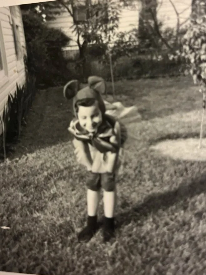 A young child dressed in a playful mouse costume, including large mouse ears, is standing on a grassy backyard lawn, smiling at the camera.