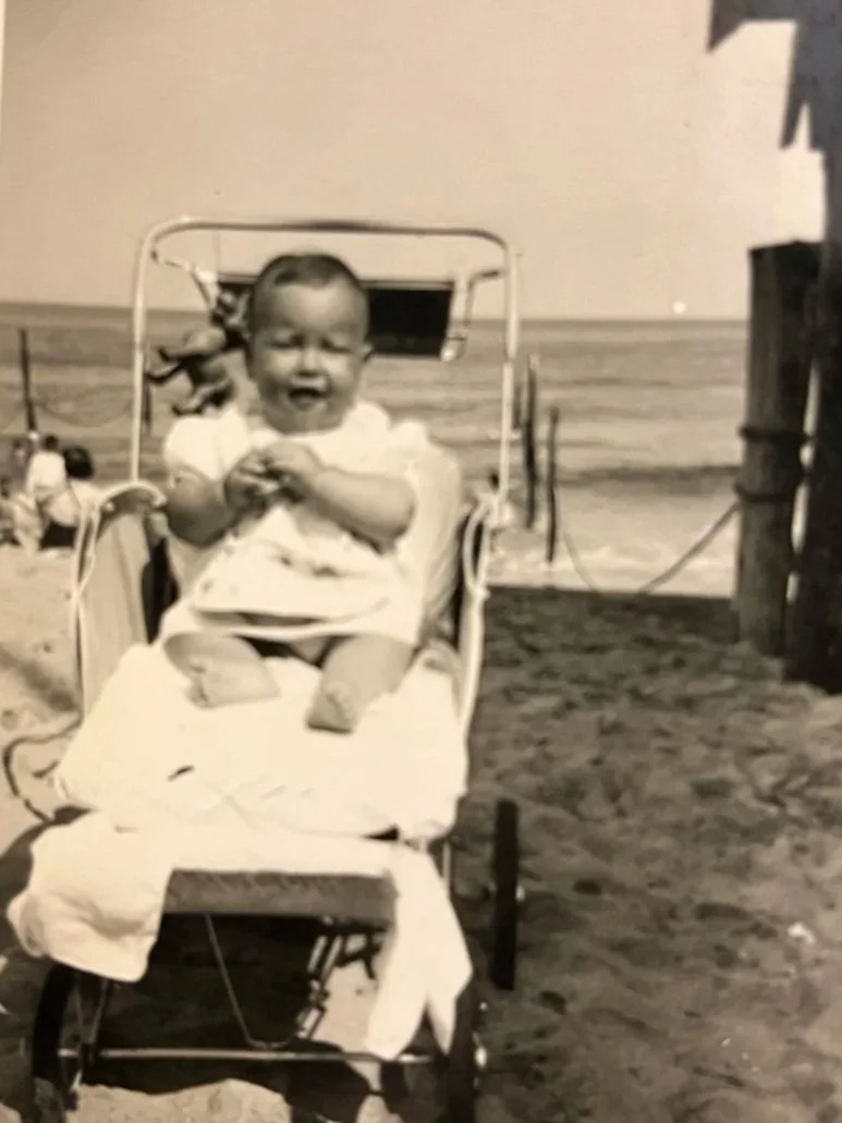 Black and white photo of a happy baby sitting in a stroller, holding an object, outdoors on a sunny day.