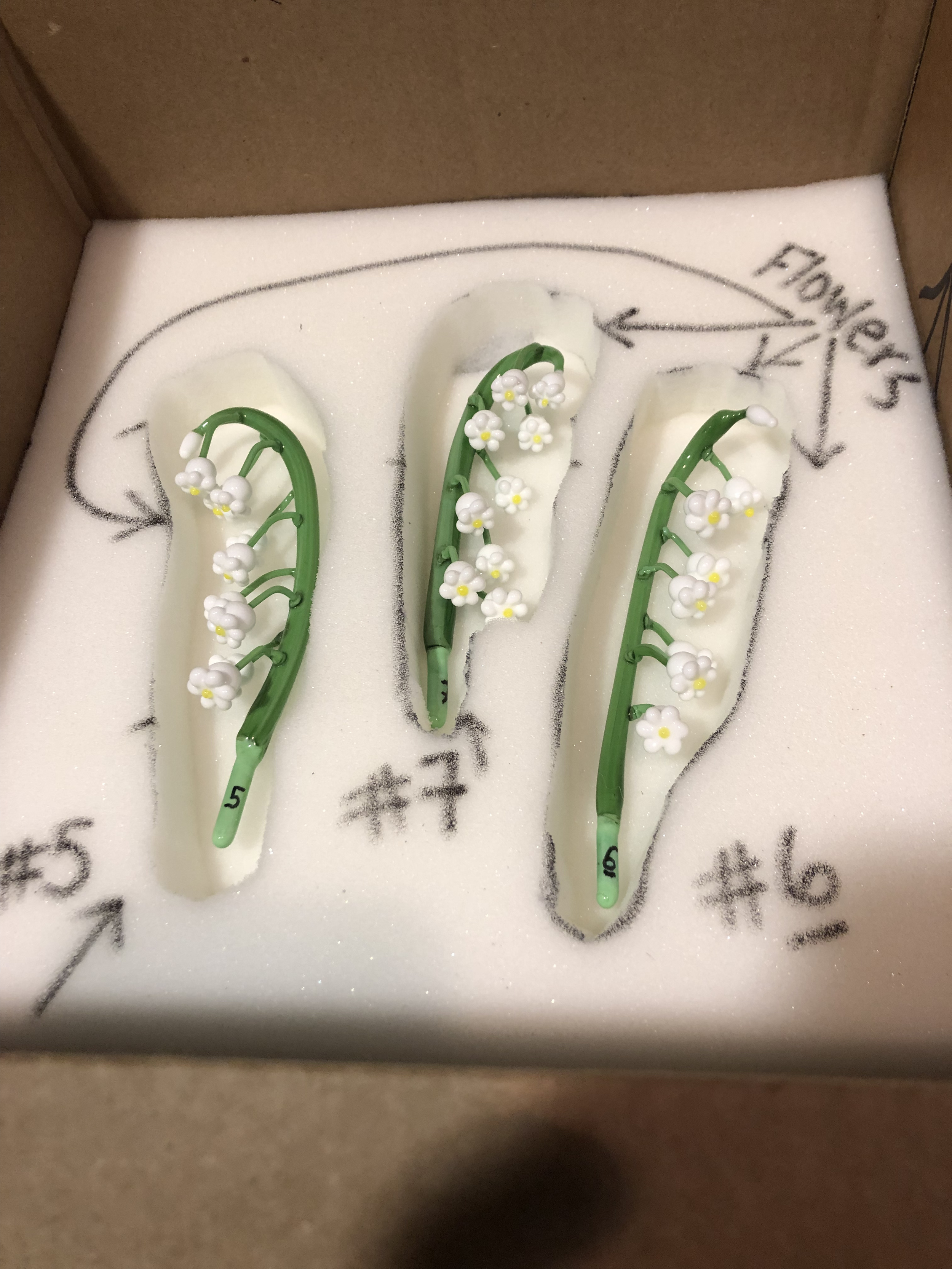 A decorative hairpin with white flowers and green stems placed inside a foam packaging with black handwritten measurements.