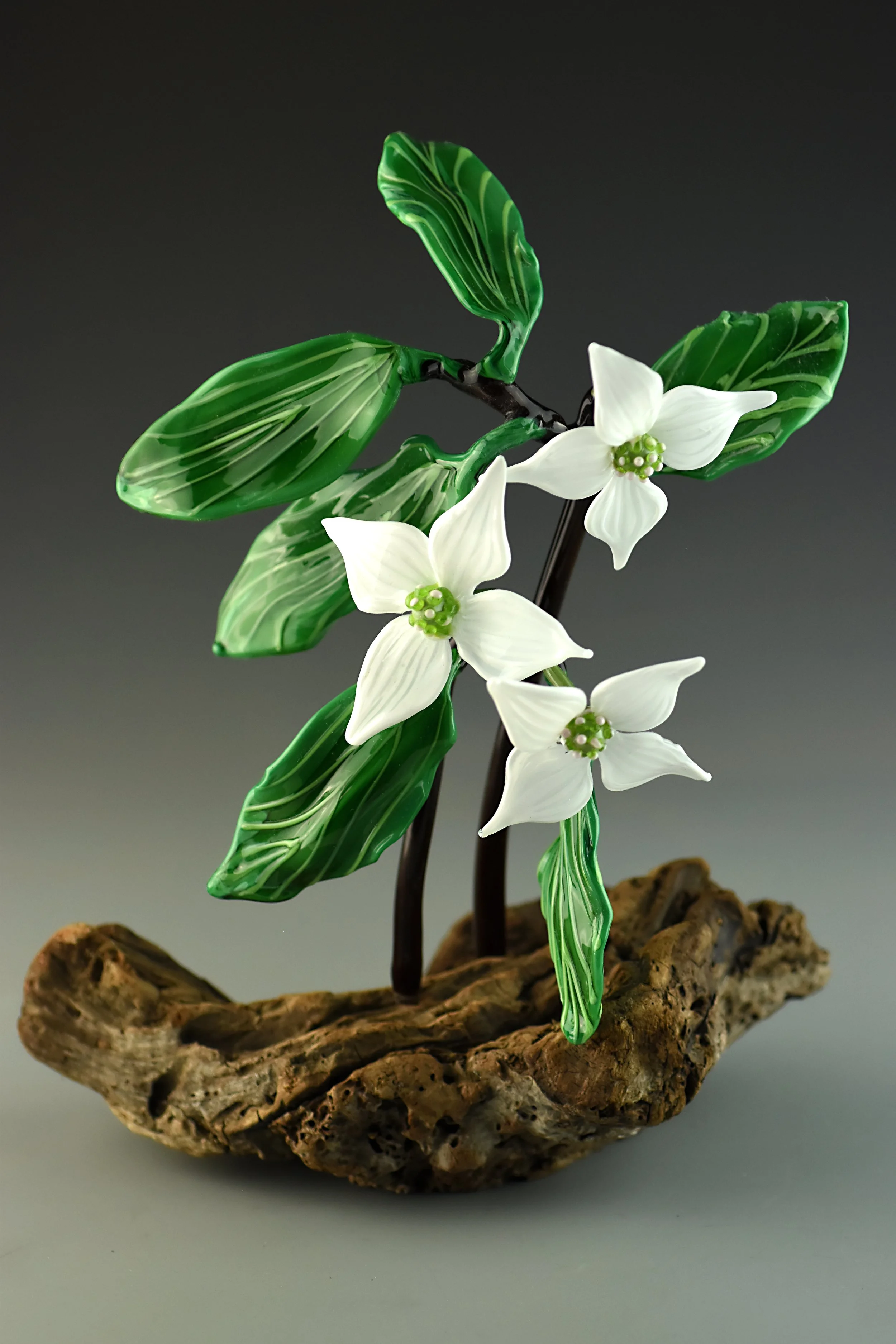 A decorative glass sculpture of white flowers and green leaves on a brown, textured base against a dark background.