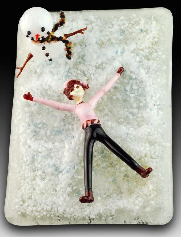 A snowman made of marshmallows with chocolate chip eyes, a small orange nose, and a twizzler scarf, along with a glass figurine of a girl in a pink sweater and black pants lying on snowy ground with arms outstretched.