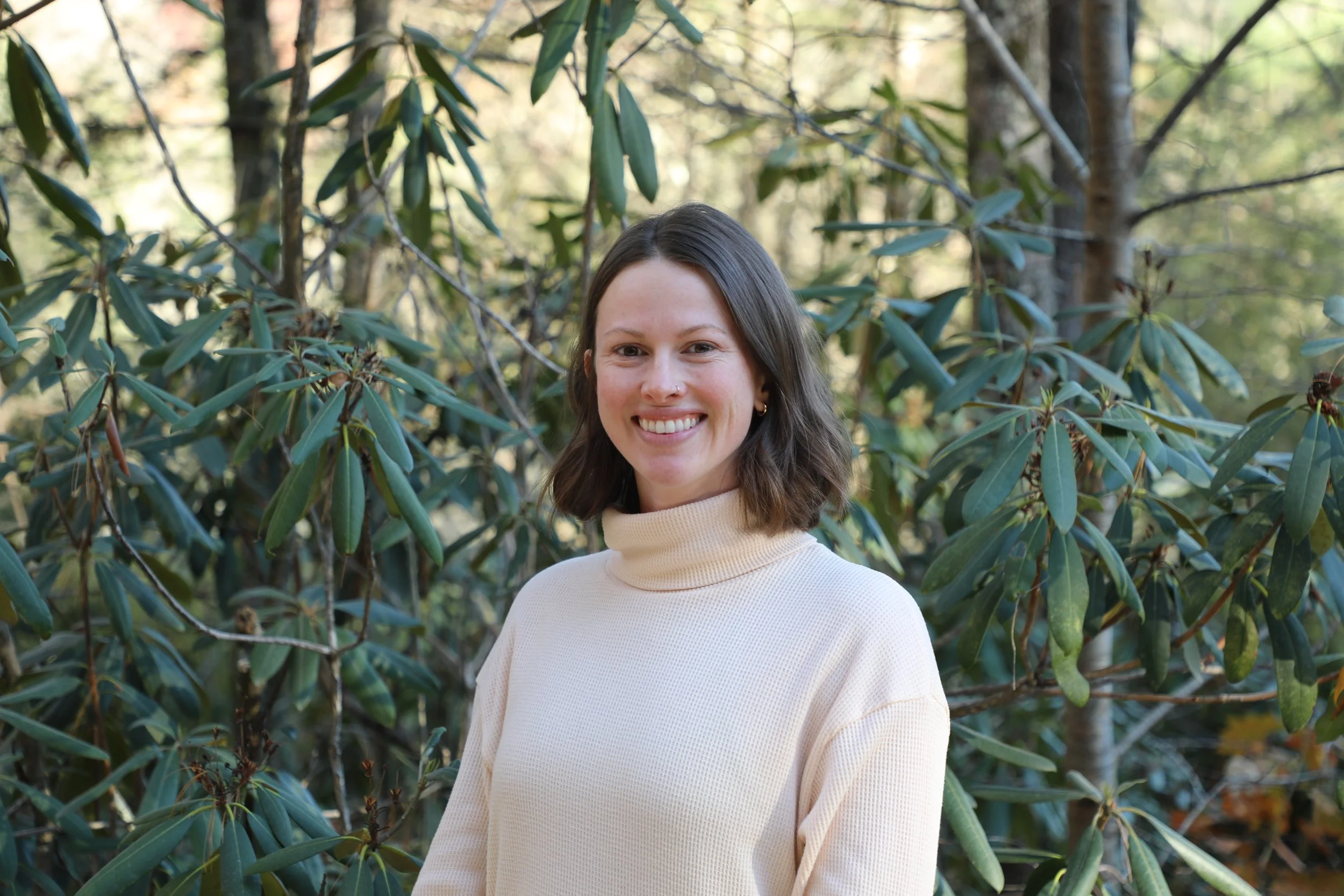 A woman with shoulder-length brown hair and a beaming smile, wearing a cream turtleneck sweater, standing amidst green foliage in a forest.