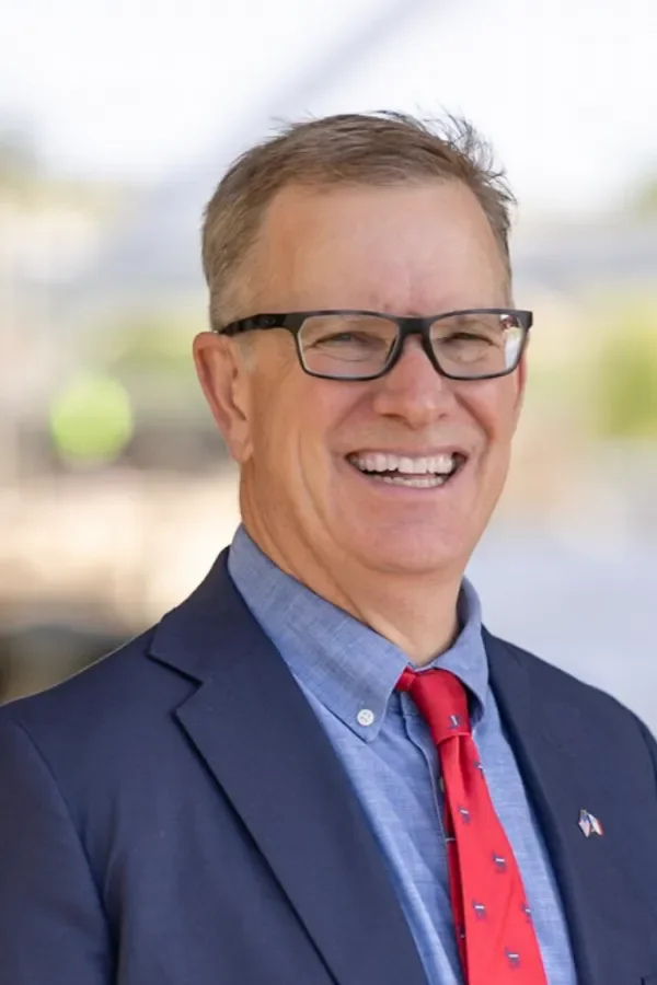 A smiling middle-aged man wearing glasses, a dark suit, a light blue shirt, and a red tie, outdoors with blurred background.