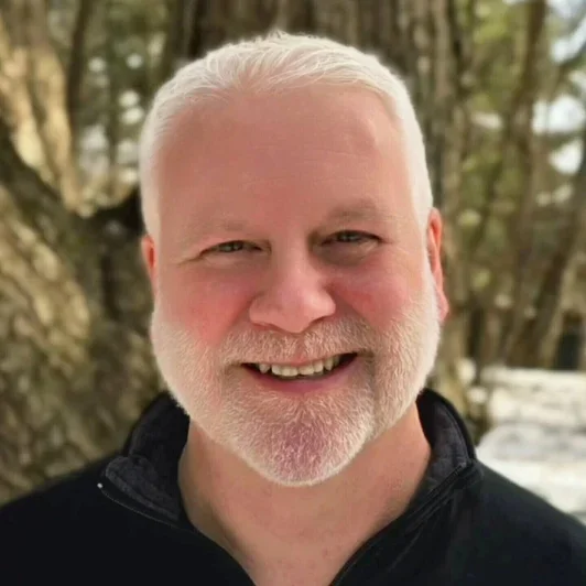 Close-up of a smiling man with white hair and beard, wearing a black jacket, standing outdoors with trees and snow in the background.