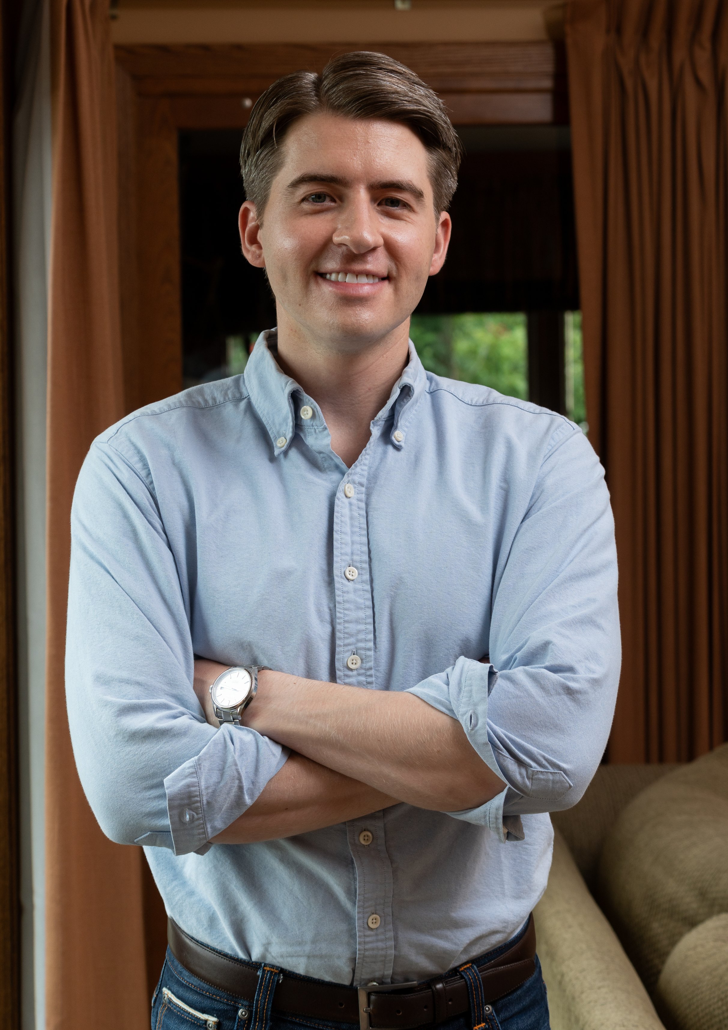A young man standing indoors, smiling with arms crossed, wearing a light blue button-up shirt, jeans, and a watch, in front of wooden walls and beige cushions.