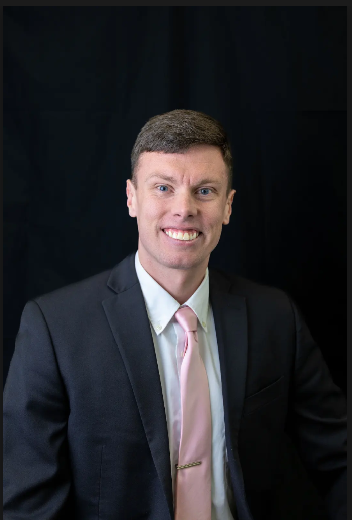 A young man in a dark suit, white shirt, and light pink tie smiling against a black background.