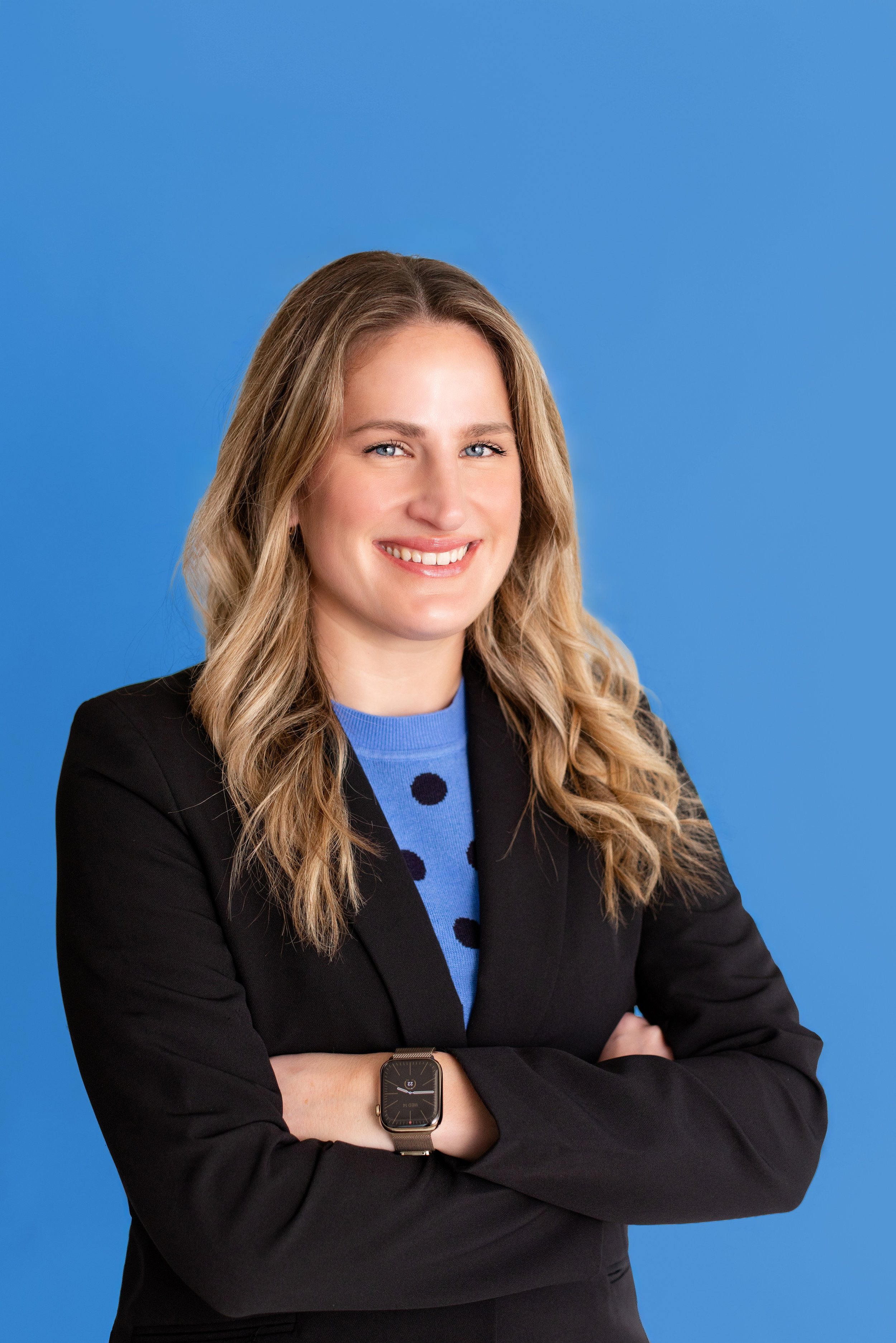 A woman with long blonde hair, wearing a black blazer over a blue polka dot sweater, smiling, with arms crossed and an Apple Watch on her left wrist, standing against a solid blue background.