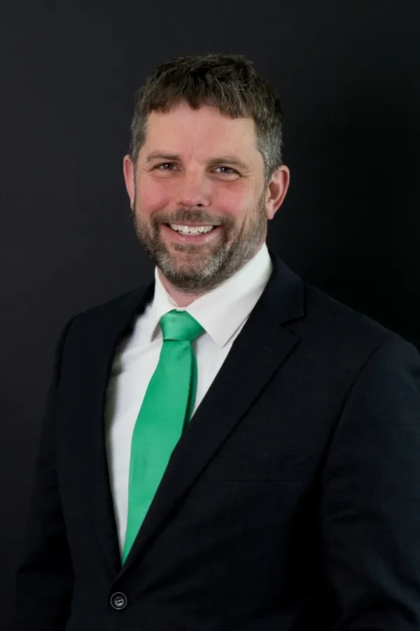A man with short dark hair, light skin, and a beard, wearing a dark suit, white shirt, and a bright green tie, smiling against a dark background.