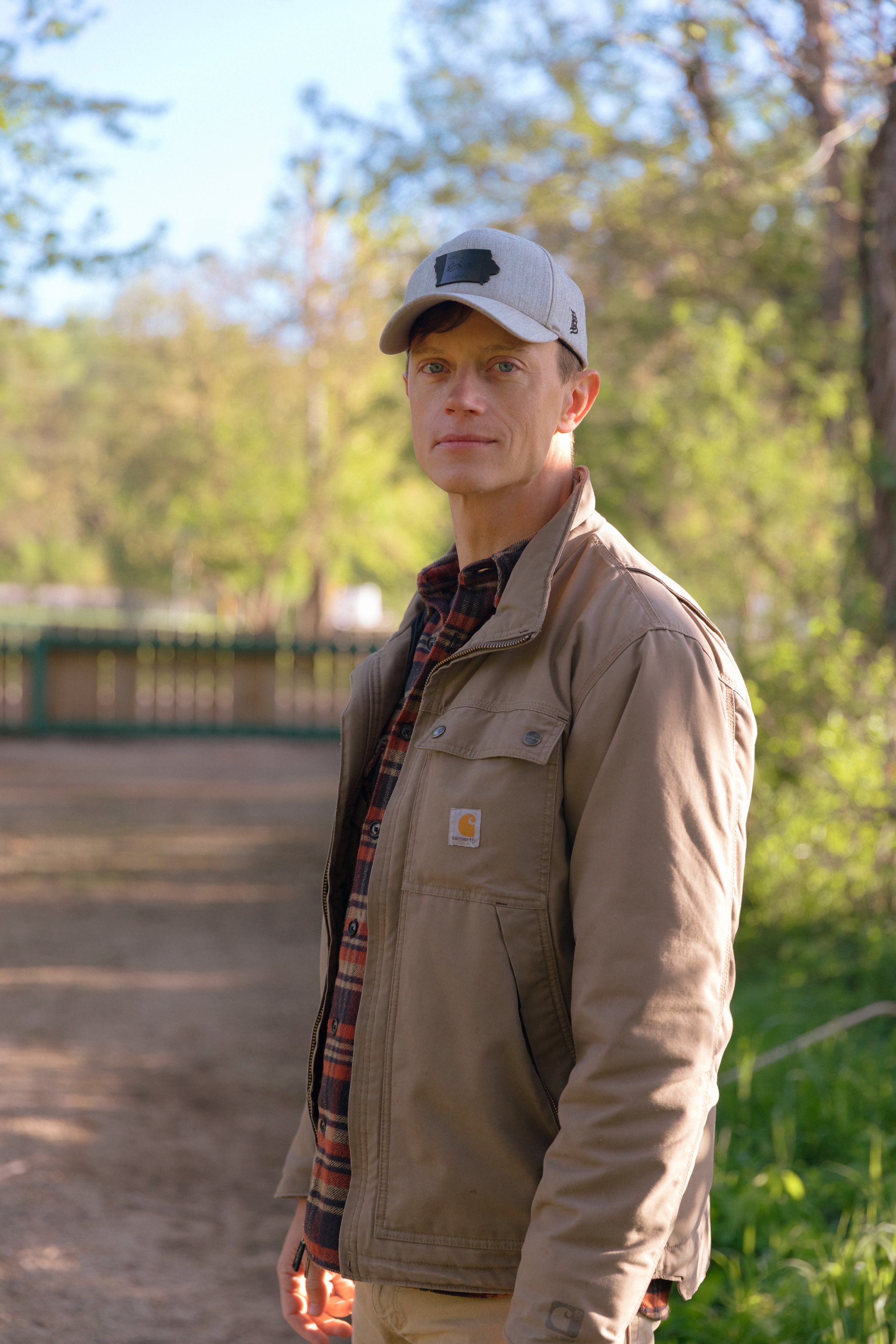 A young man wearing a gray baseball cap, beige Carhartt jacket, and plaid shirt standing outdoors in a park or natural area with trees and greenery in the background.