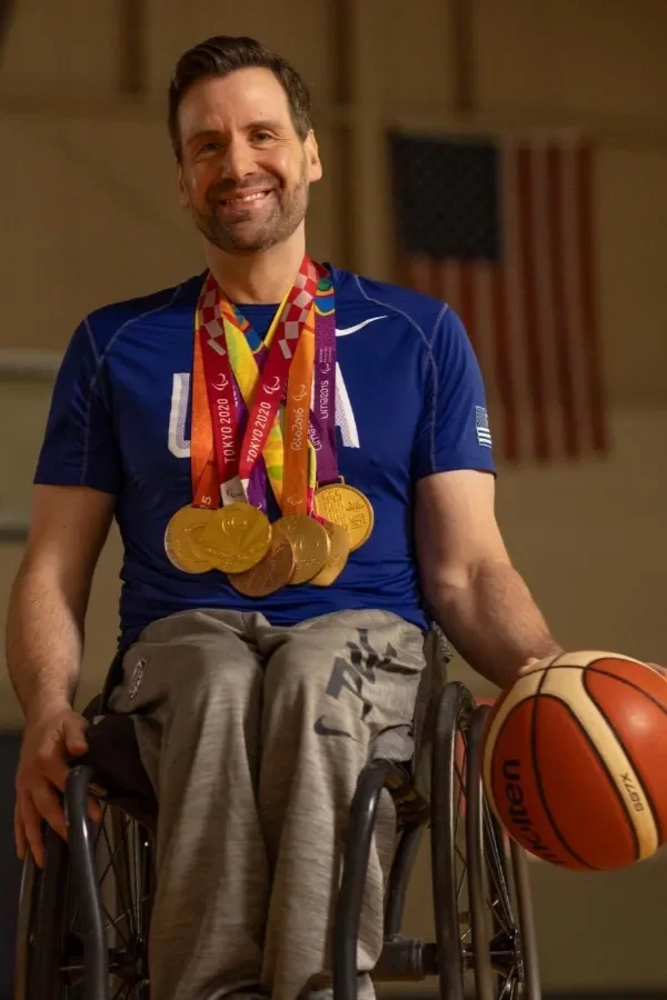 A man in a wheelchair wearing a blue sports shirt with multiple gold medals around his neck, holding a basketball, and smiling.