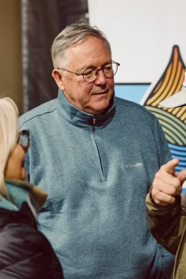 An older man with glasses and gray hair wearing a blue quarter-zip fleece jacket, engaged in conversation with two women, one with blonde hair and glasses, in an indoor setting.