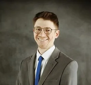 A smiling man in a gray suit, white shirt, and blue tie, wearing glasses, in front of a gray background.