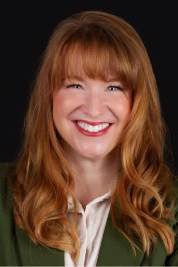 A woman with long red hair, bright smile, white shirt, and green blazer against a black background.