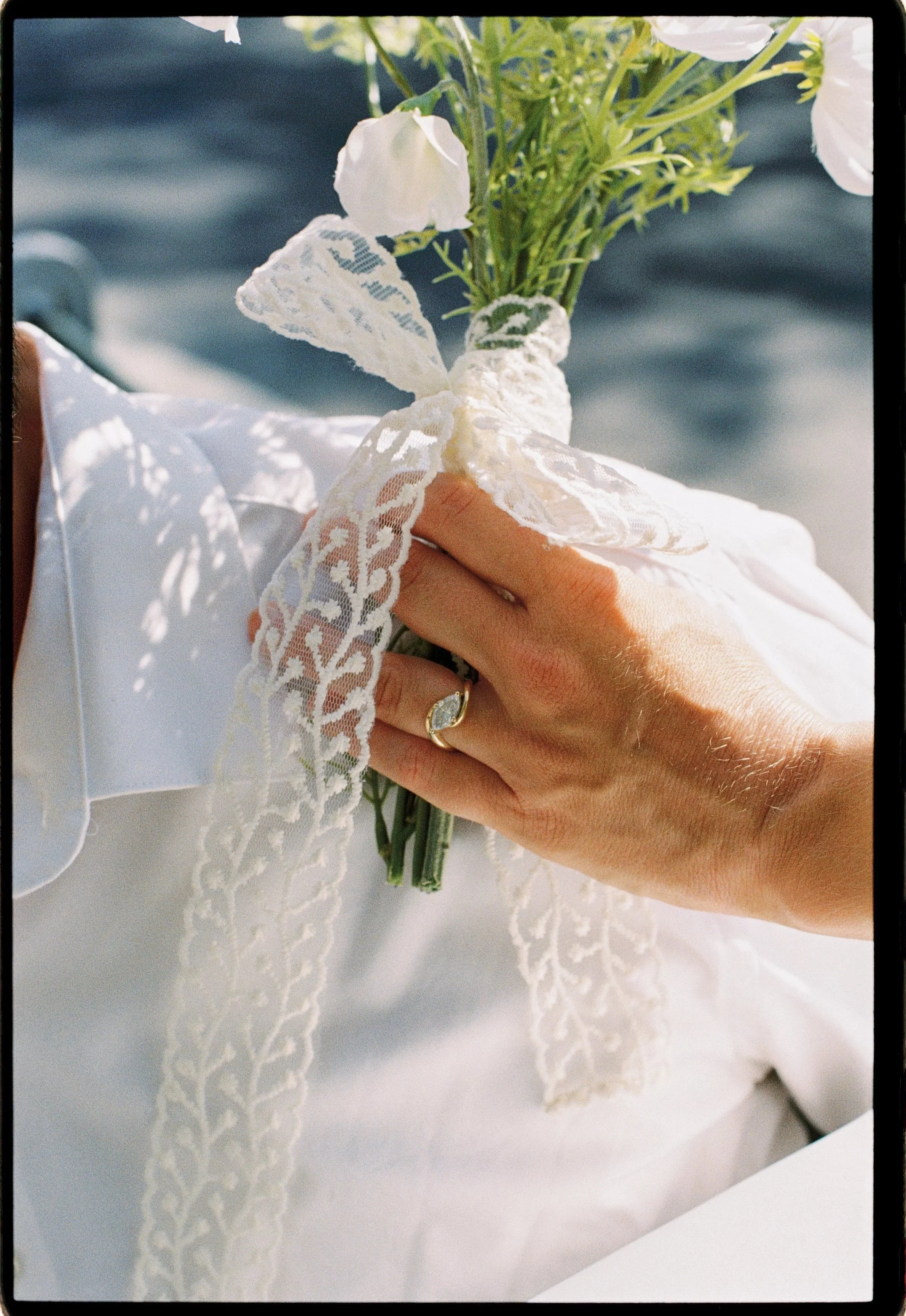Couples and engagement film photography in Paris. A hand holding a small bouquet of white flowers wrapped in lace fabric, with a white lace ribbon tied around the stems, on a white fabric surface with lace embroidery.
