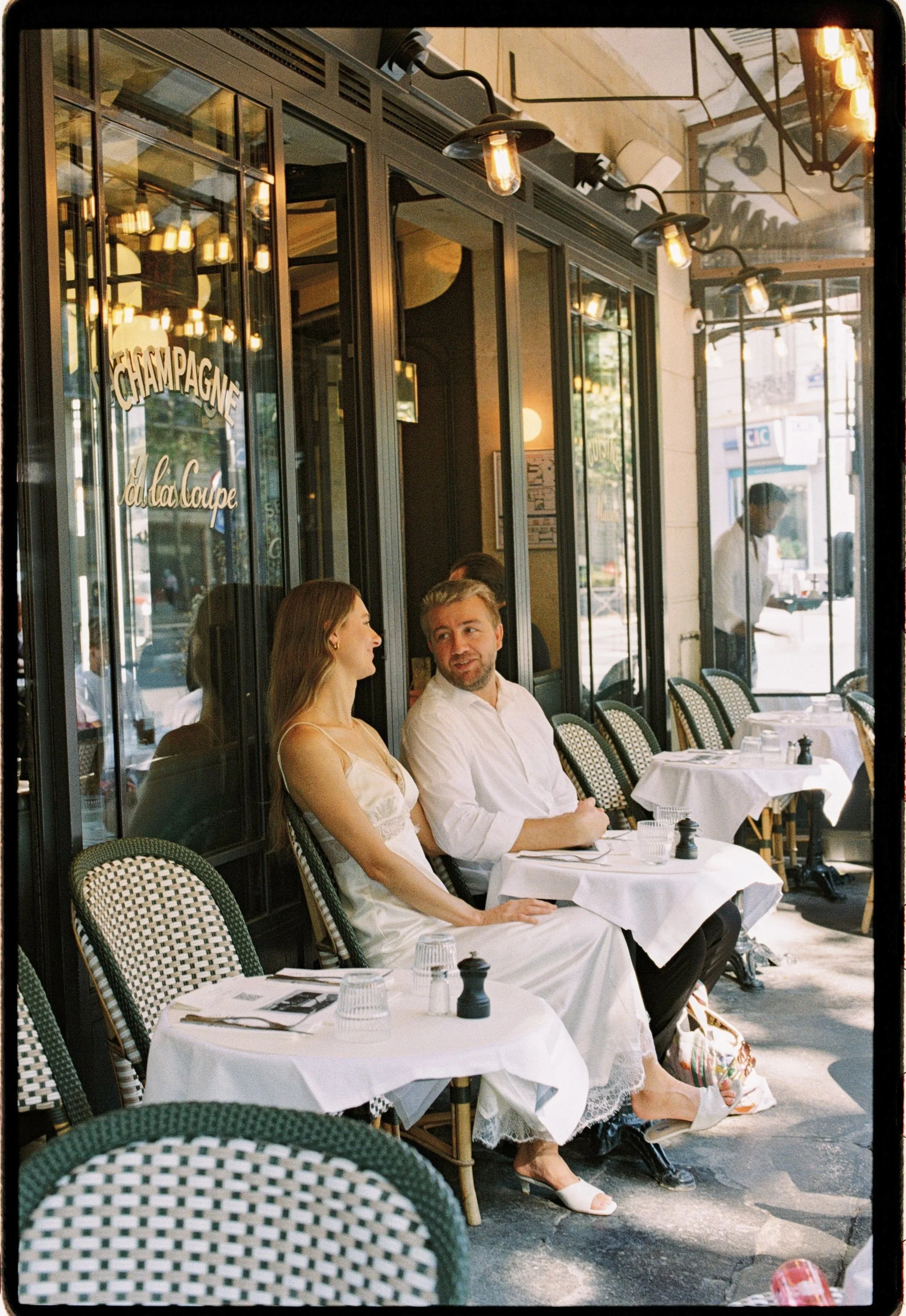 	
Couples and engagement film photography in Paris.Two people sitting and talking at a dining table outside a Parisian bistro. The woman is wearing a white dress, and the man is in a white shirt. The table has white tablecloths, glasses, and cutlery.