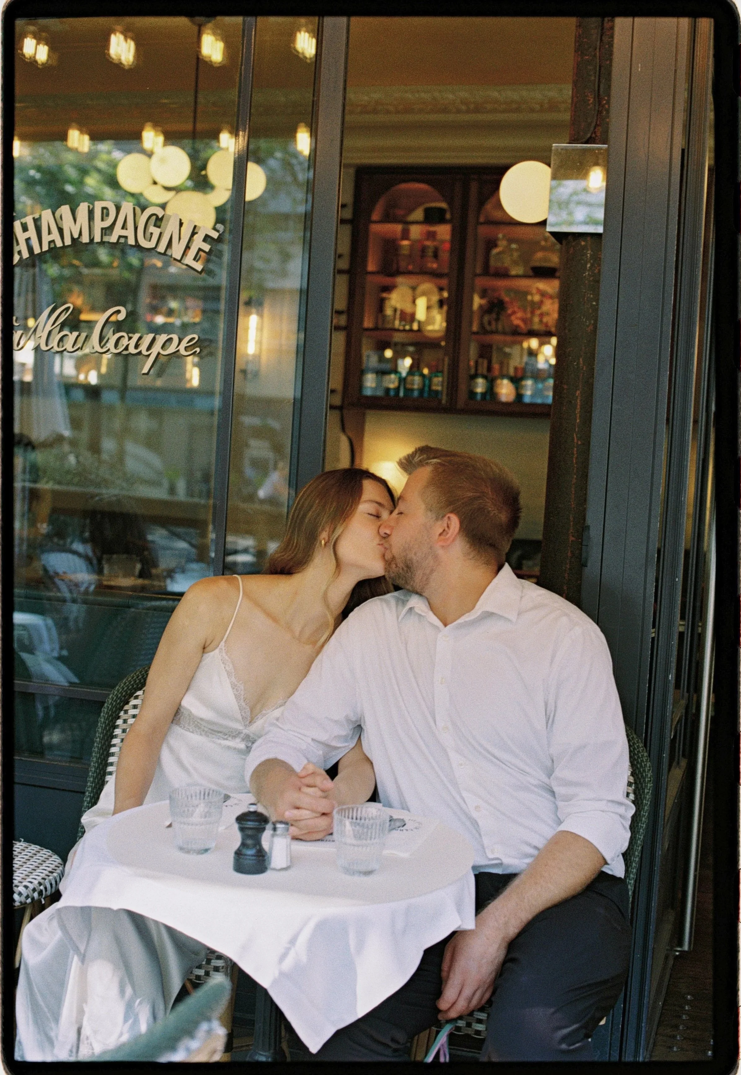 A young woman and man are kissing at a table in a restaurant. The woman has long hair and is wearing a white dress. The man has short hair and a beard, wearing a white shirt. They are holding hands on the table, which has glasses of water, salt and p