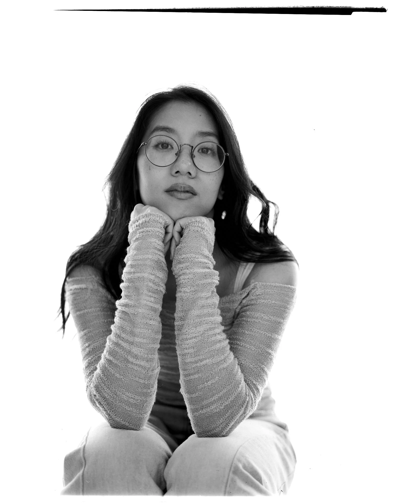 Black and white portrait of a young woman with glasses and long hair, sitting with her chin resting on her hands, wearing a sweater with fingerless gloves.