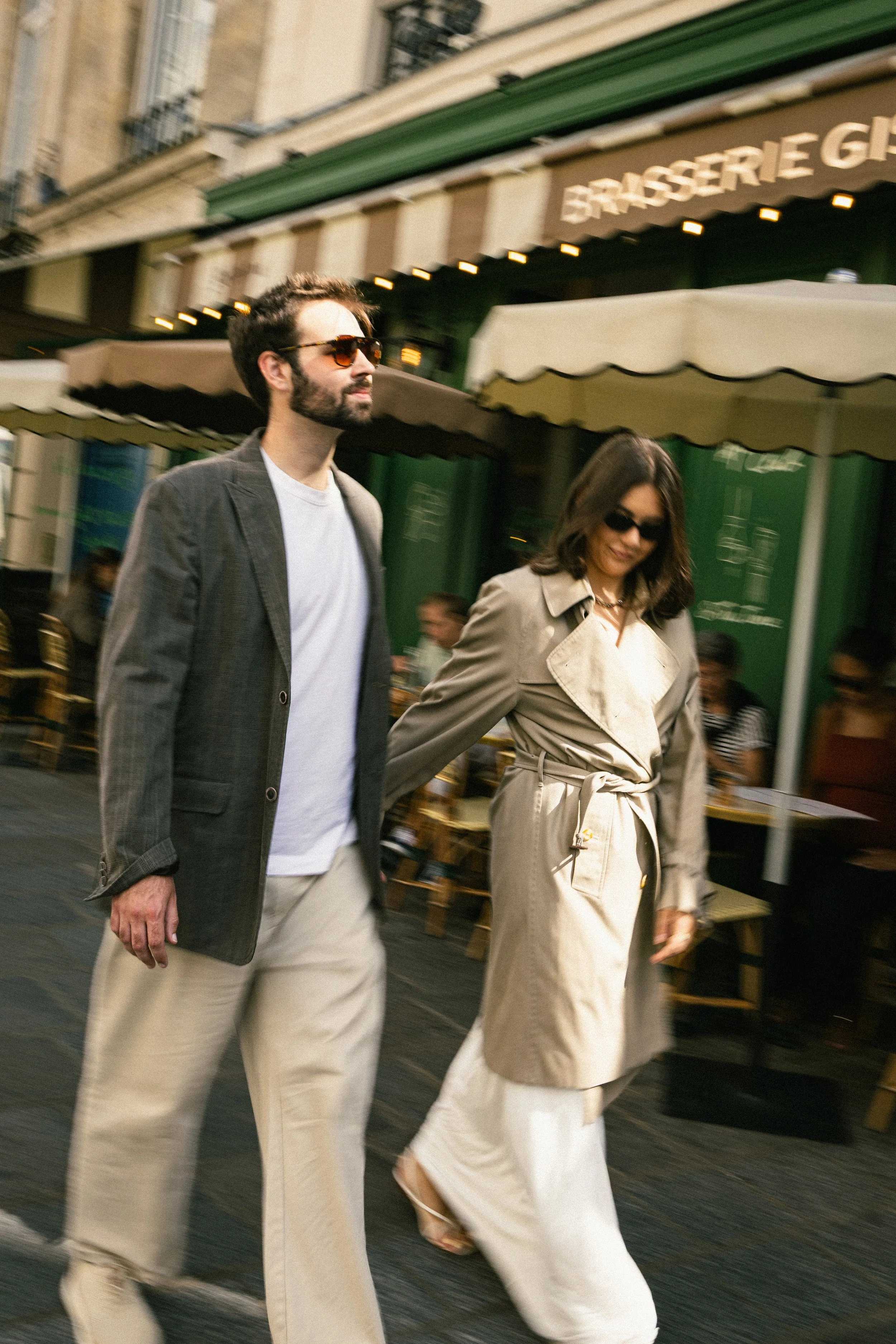 Two stylish people walking on a city street, wearing sunglasses and beige clothing, with a café and outdoor seating in the background.