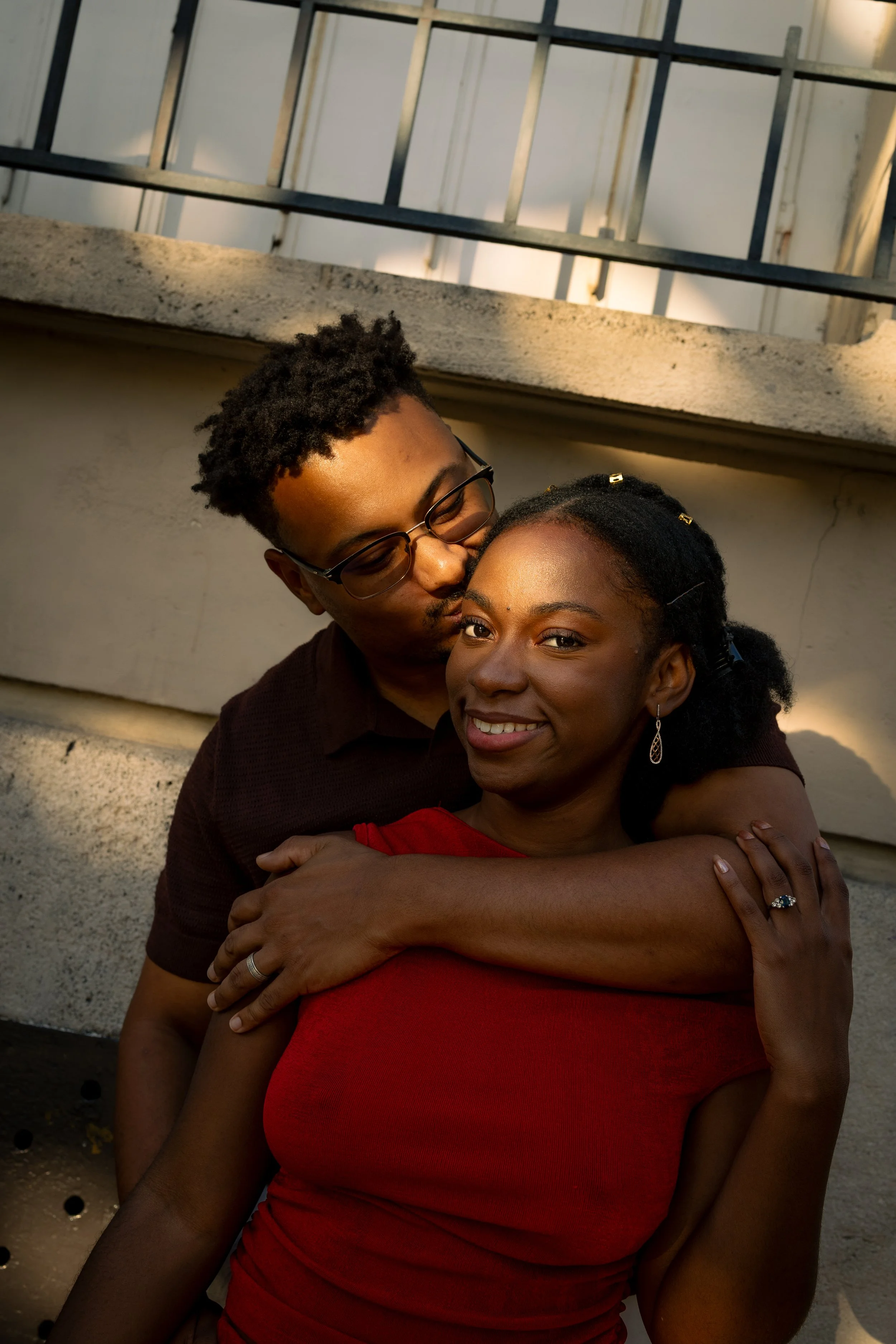 Couples and engagement film photography in Paris. A couple hugging outdoors, with a woman in a red dress and a man wearing glasses and a dark shirt, standing near a concrete wall and a metal staircase.